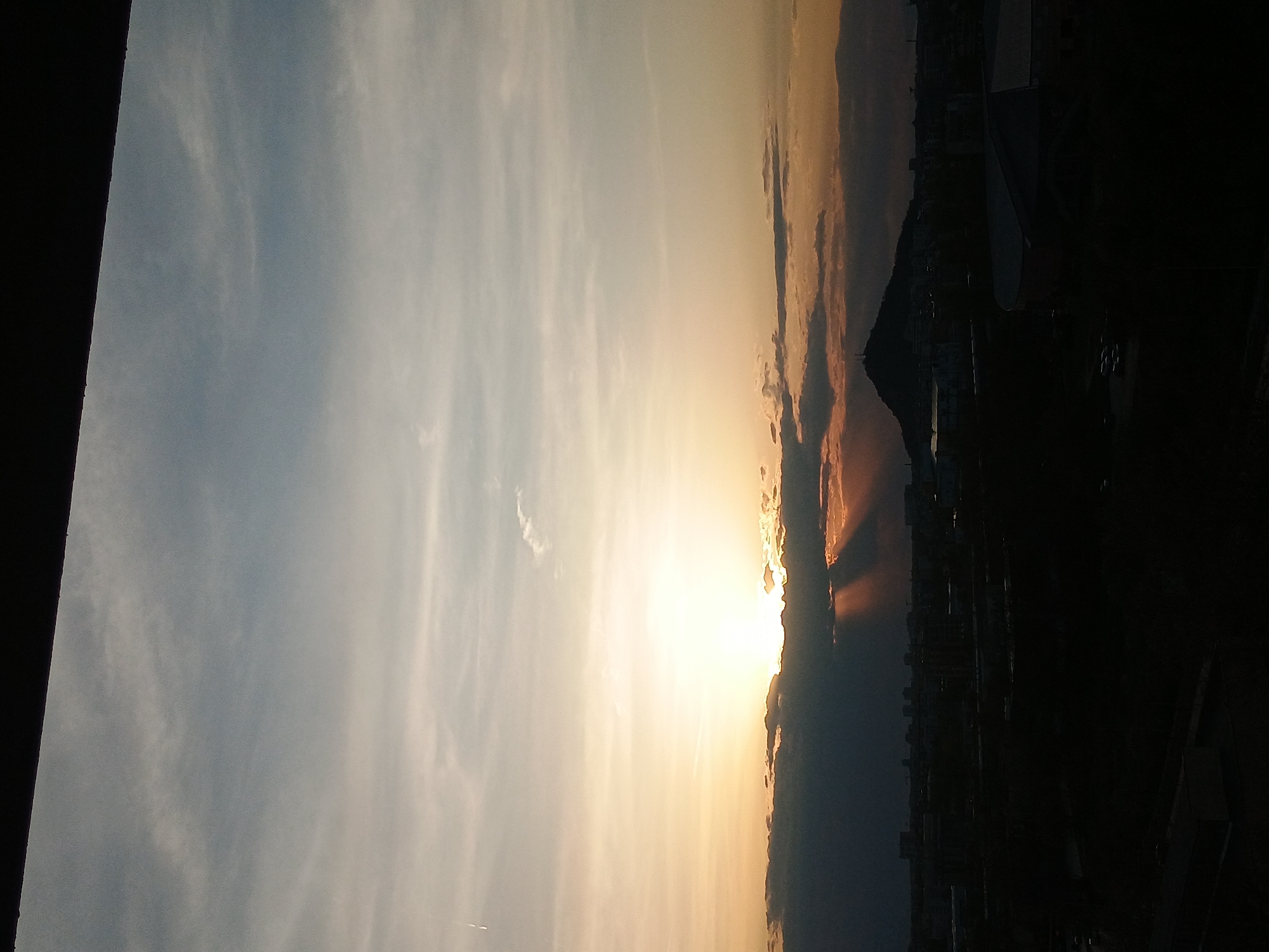 The image depicts a sunset over Plovdiv, Bulgaria, with the setting sun obscured by clouds on the horizon. Sunbeams are visible. The silhouette of the city skyline, including a hill or mountain (likely one of the hills of Plovdiv), is visible in the foreground. The left side of the frame shows a dark, vertical structure. The sky progresses from blue on the left to orange and yellow near the horizon.