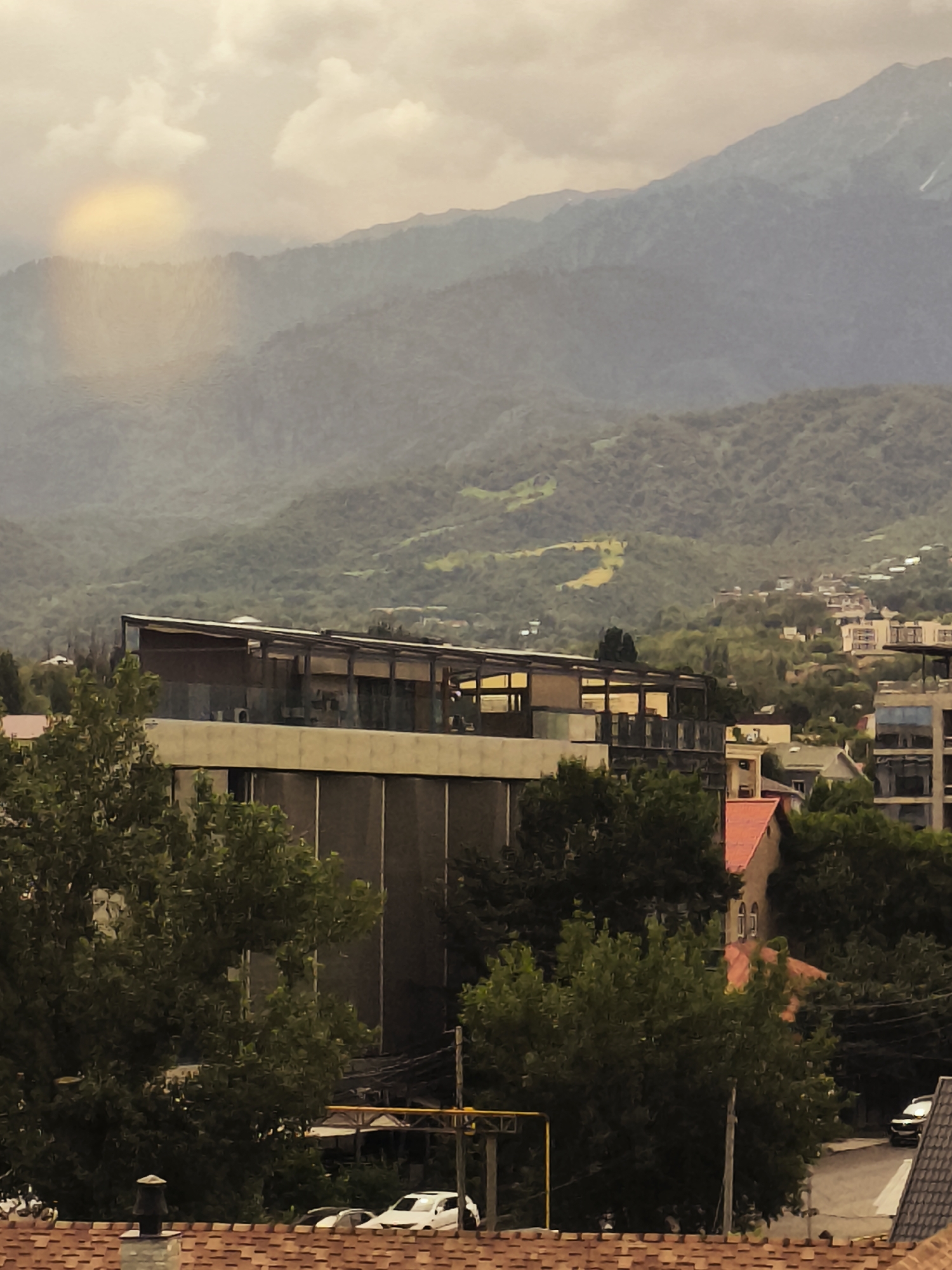 The image captures a panoramic view of the city of Almaty, Kazakhstan, with a backdrop of majestic mountains. In the foreground, a building with a modern architectural design, featuring a flat roof and large windows, is prominent. Trees with lush green foliage surround the building, adding a natural element to the urban landscape. The mountains, covered in a dense forest, rise majestically in the background, their peaks partially obscured by a light mist. The sky above is overcast, with clouds casting a soft, diffused light over the scene. A halo-like effect is visible in the sky, possibly due to atmospheric conditions or a lens flare. The city's architecture, a blend of modern and traditional, is visible in the mid-ground, with residential buildings nestled amidst the greenery. The overall impression is one of a serene and picturesque city, where urban life harmoniously coexists with the grandeur of nature.