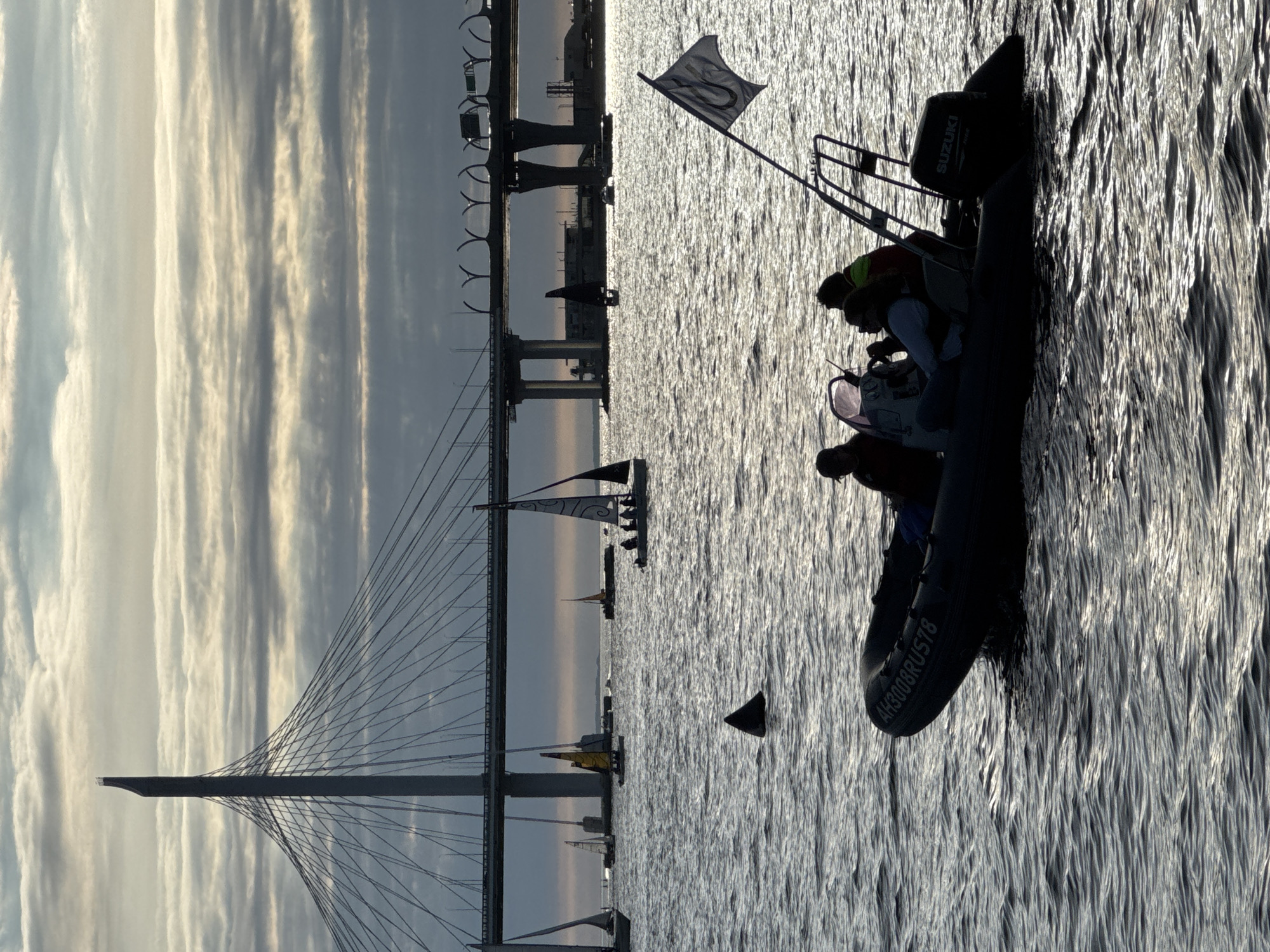 The image captures a panoramic view of the Neva River in Saint Petersburg, Russia, under a dramatic, cloud-filled sky. Dominating the background is the Western High-Speed Diameter, a modern cable-stayed bridge, with its distinctive tower and suspended roadway silhouetted against the sky.
In the foreground, a rigid inflatable boat (RIB) carrying four individuals is navigating the choppy waters. The boat is dark and appears to have the registration "AH3008RUST8" visible on its side. The individuals are silhouetted, making their features indistinct, but they appear to be casually dressed, some wearing life vests. One person is at the helm, steering the boat. The boat's Suzuki outboard motor is visible at the stern.
Scattered across the water are several sailboats of varying sizes. Some are closer to the foreground, while others are further away, near the bridge. The sails are either furled or partially raised, suggesting the boats are either preparing to sail, returning to shore, or taking part in a regatta.
A notable detail in the mid-ground is the dark triangular fin of what appears to be a shark breaking the surface of the water. This is an unusual and attention-grabbing element within the scene.
The overall atmosphere of the image is one of a late afternoon or early evening, with soft, diffused light filtering through the clouds, casting a slightly muted, atmospheric glow on the water and the structures. The texture of the water is characterized by small, rippling waves, reflecting the overcast sky. The scene combines elements of urban infrastructure, recreational boating, and a touch of the unexpected with the shark's fin.