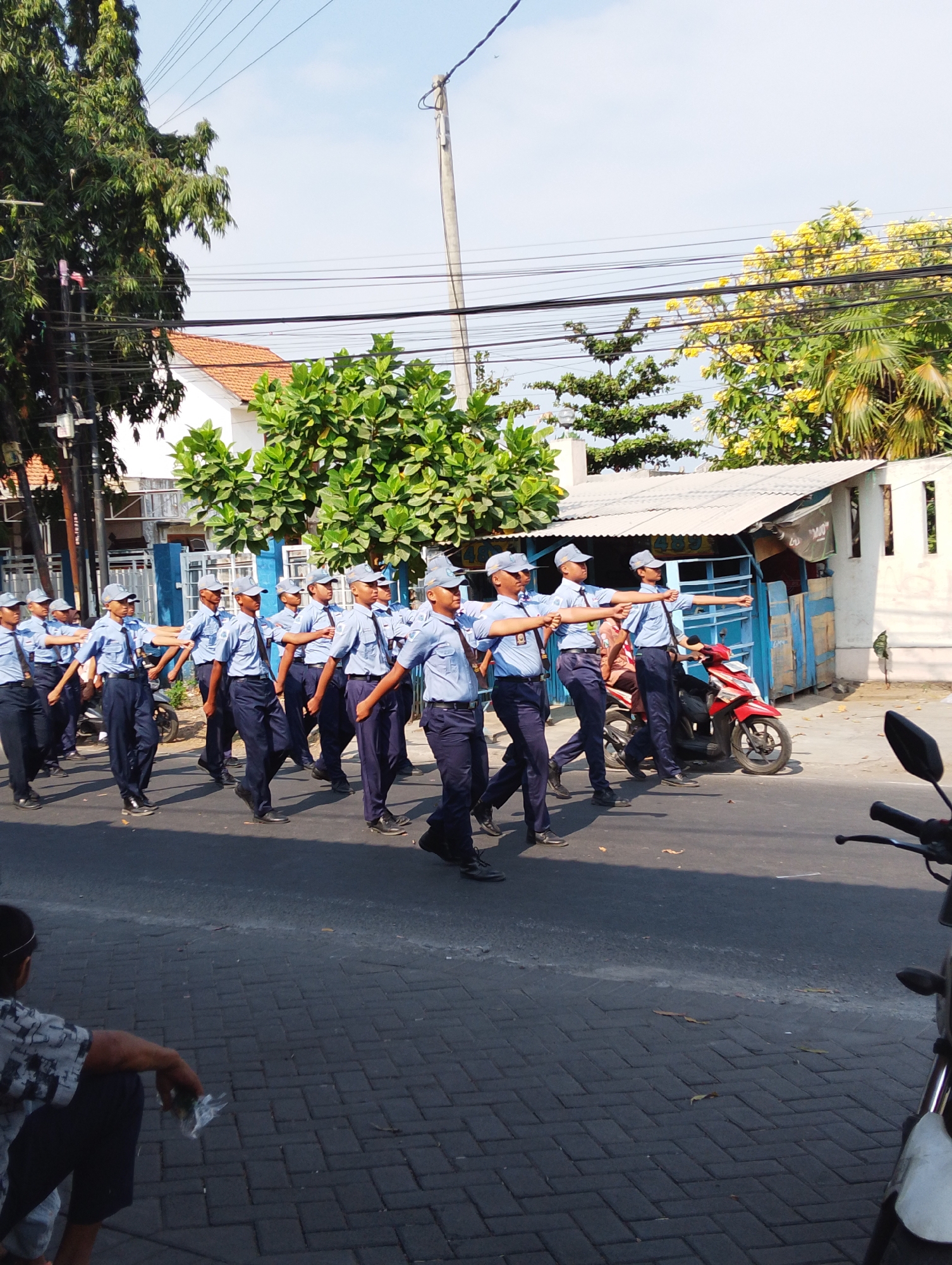 In Manaruwi, Indonesia, a group of young people in matching blue uniforms and caps are marching in formation on a paved road. They appear to be participating in a parade or drill, with their arms extended to the side in unison. The background features a residential area with houses, trees, and power lines overhead. A red motorcycle is parked to the right of the marching group. A person is seated on the left side of the road, observing the procession. The scene is set under a clear blue sky, suggesting a sunny day.