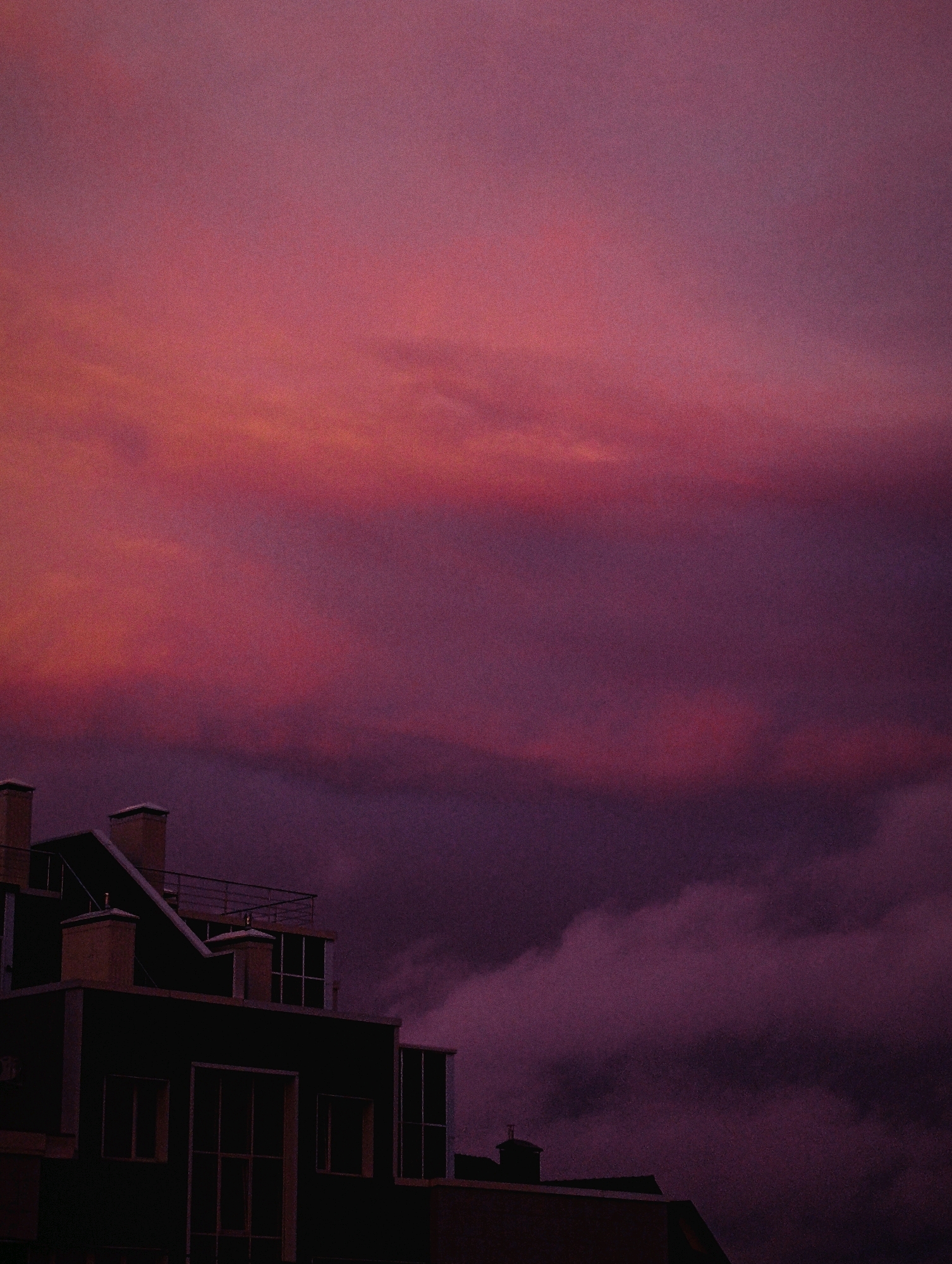This is a medium shot, low-angle photograph of a dark building against a vibrant, dusky sky. The building is dark, with visible windows and chimney-like structures on its roof. The sky is filled with soft, diffused clouds that range in color from deep purple to bright pink and orange, suggesting sunset or sunrise. The overall mood of the image is atmospheric and somewhat dramatic due to the intense colors of the sky. The location is described as Unknown, Unknown.