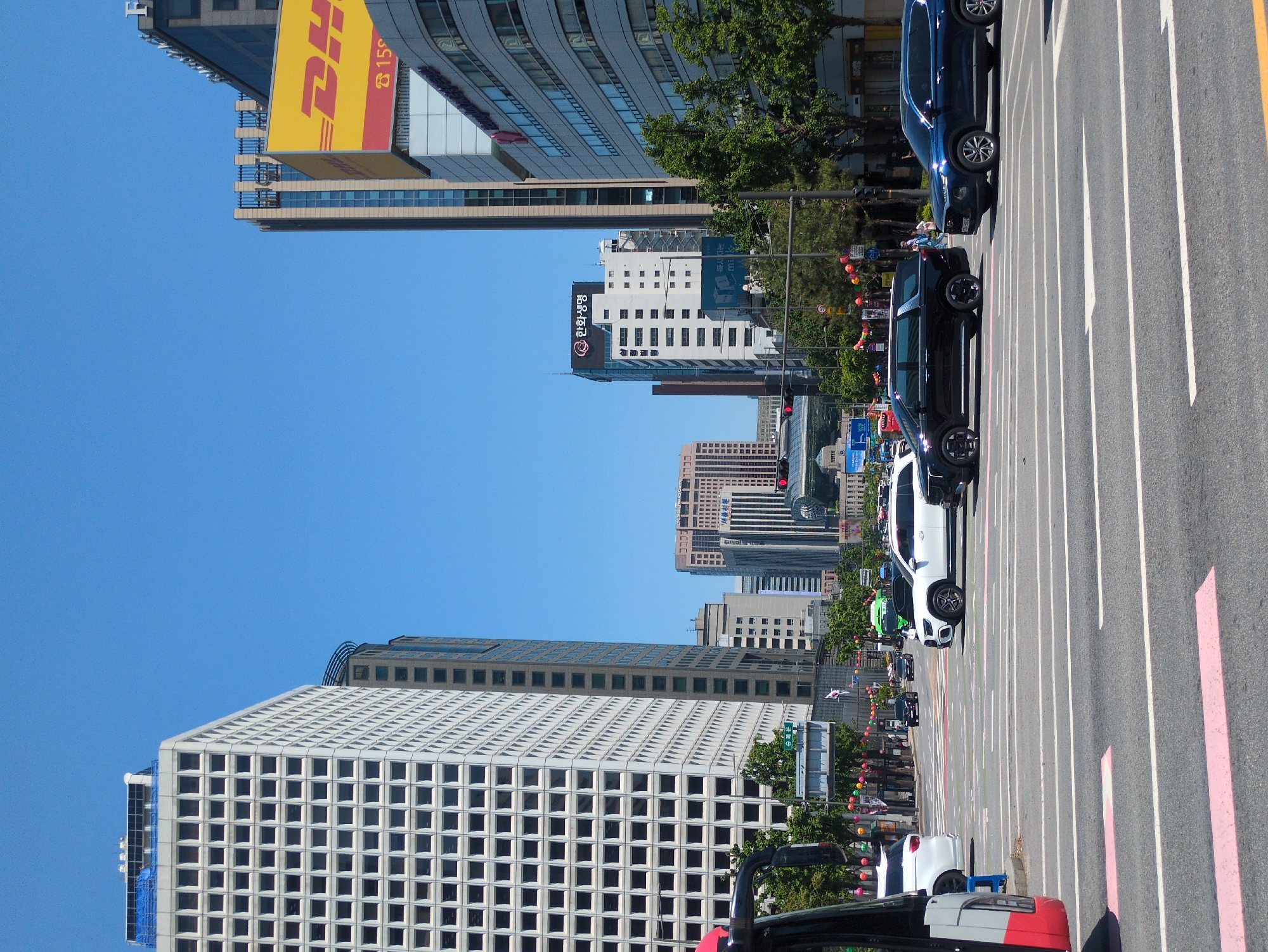 The image shows a street in Seoul, South Korea, with a view of several tall buildings. On the left is a large white building with a grid-like facade. Further down the street, a DHL sign is visible on another tall building. Other skyscrapers are visible in the background. Cars are parked along the right side of the street. The sky is clear and blue. Trees line the street, and colorful lanterns are strung between the trees. The image is oriented vertically, with the buildings appearing to stand on their sides.