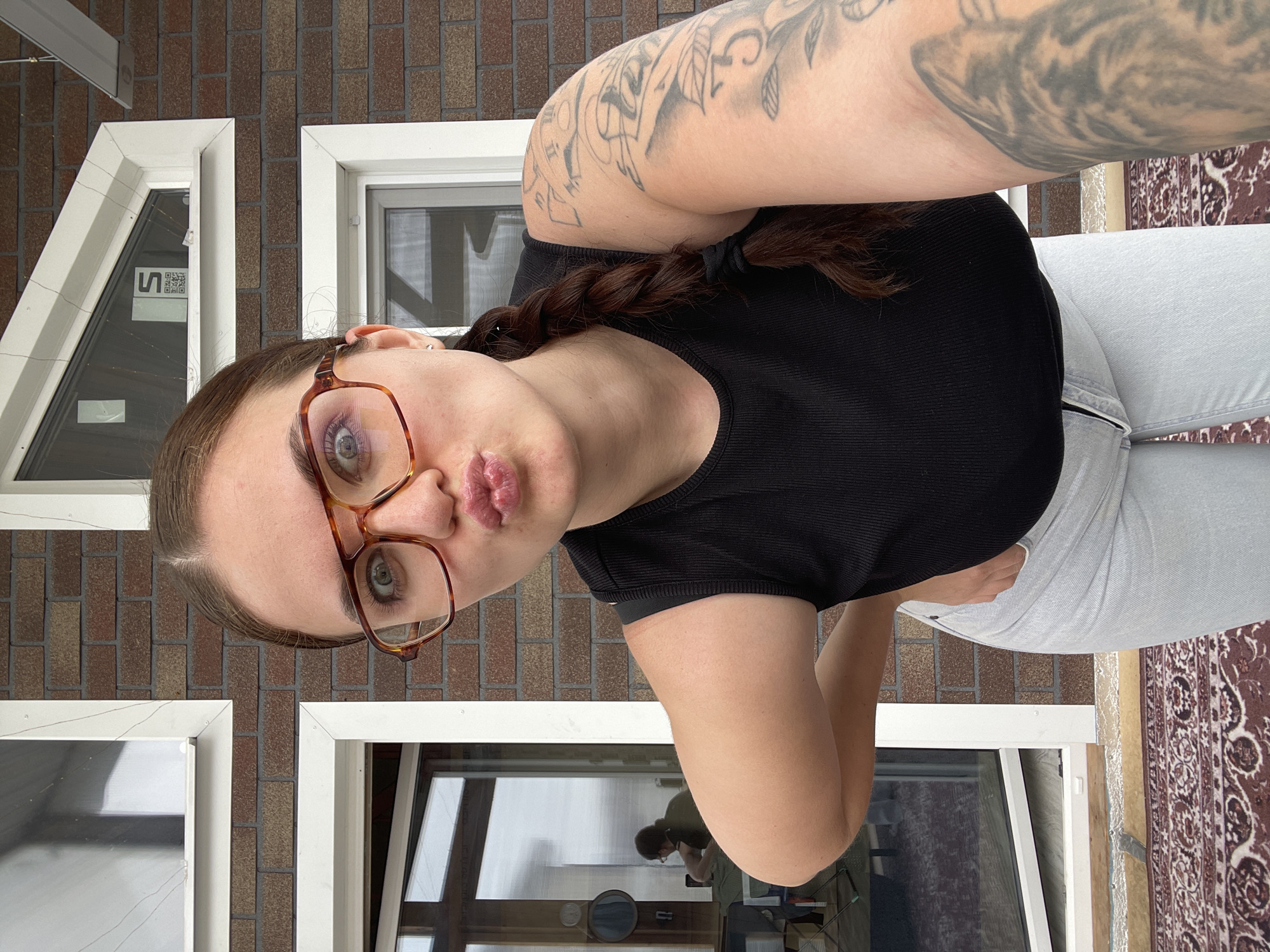 A young woman is taking a selfie outdoors. She is wearing glasses, a black top, and light-colored jeans. She has a braided hairstyle and tattoos on her arm. The background shows a brick wall with white-framed windows and a brown patterned rug. The location is identifiable as Мосметаллоконструкция Онт, Russia, based on the presence of distinctive architectural features.