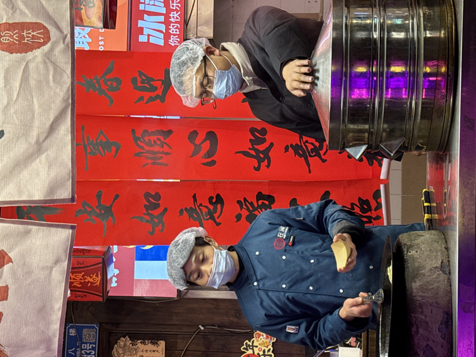 Two adult males are engaged in food preparation at a brightly lit indoor food stall in Yuzhong District, China. The man on the left, wearing a dark blue chef's jacket with a logo, a medical mask, and a hairnet, holds a small, light-colored, boat-shaped container in his left hand and a metal utensil over a dark, circular cooking surface with his right. The man on the right, wearing a dark jacket, eyeglasses, a medical mask, and a hairnet, rests his right hand on the domed lid of a three-tiered stainless steel steamer. The foreground features a reflective metallic counter, showing purple light reflections, with yellow and black tools placed between the individuals. The background is dominated by multiple vertical red banners featuring black Chinese calligraphy, including characters that translate to wishes for smooth affairs and happiness. A blue sign with white characters "八楼 3号" (Eighth Floor, No. 3) is visible on the upper left. A partial sign on the far right displays blue text, including "冰" (ice) and "你的快乐" (your happiness).