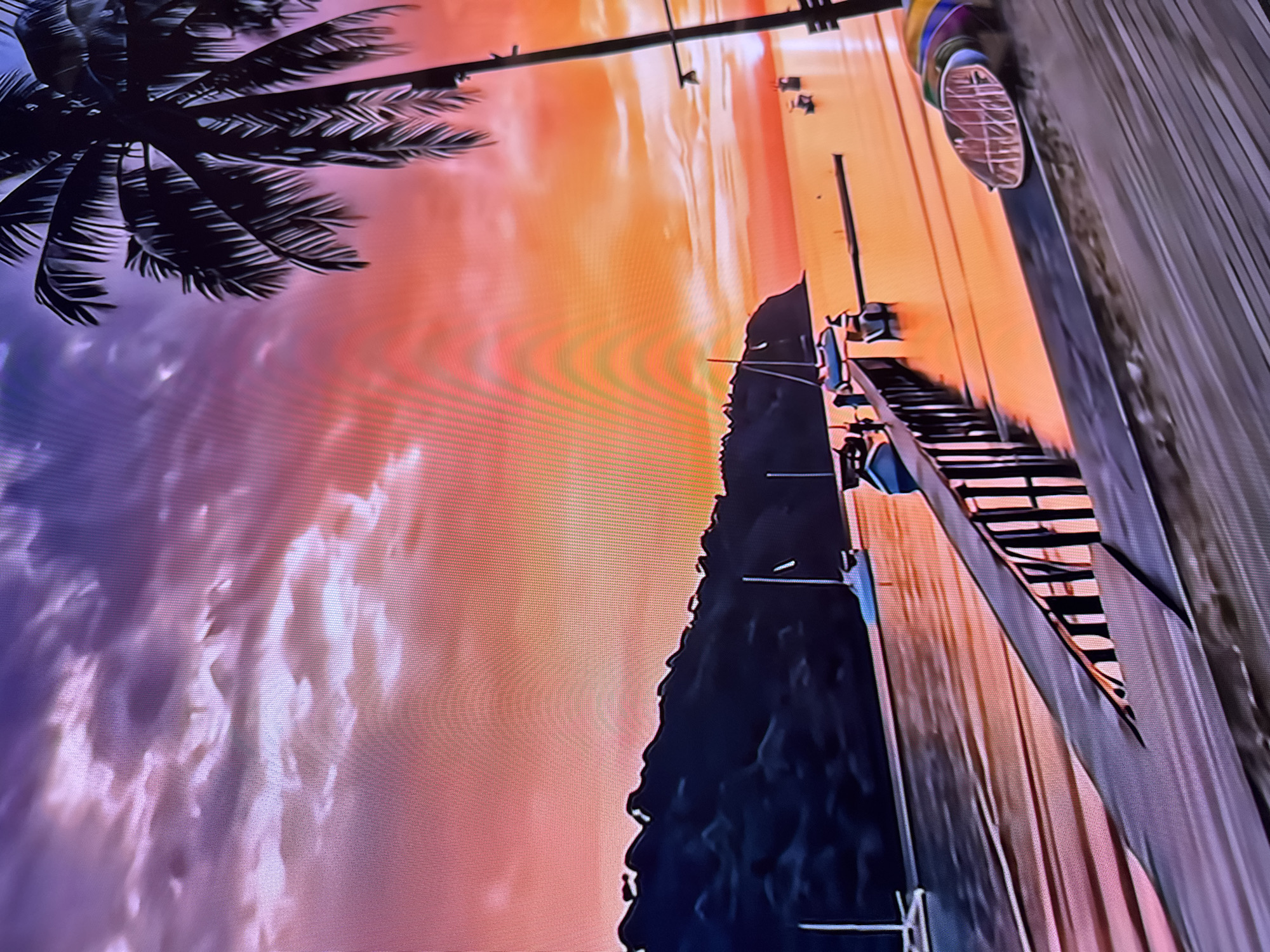 The image depicts a stylized coastal scene at sunset. A palm tree is visible in the upper left corner. The sky transitions from purple to orange hues, suggesting either sunrise or sunset. In the distance, several figures are engaged in beach activities along the shoreline. A dark, mountainous landmass is situated between the sky and the beach. A pier extends into the water, with several people standing or walking along it. There is a road with a vehicle parked on the right of the pier. This scene doesn’t feature locations of Sofia, Bulgaria.
