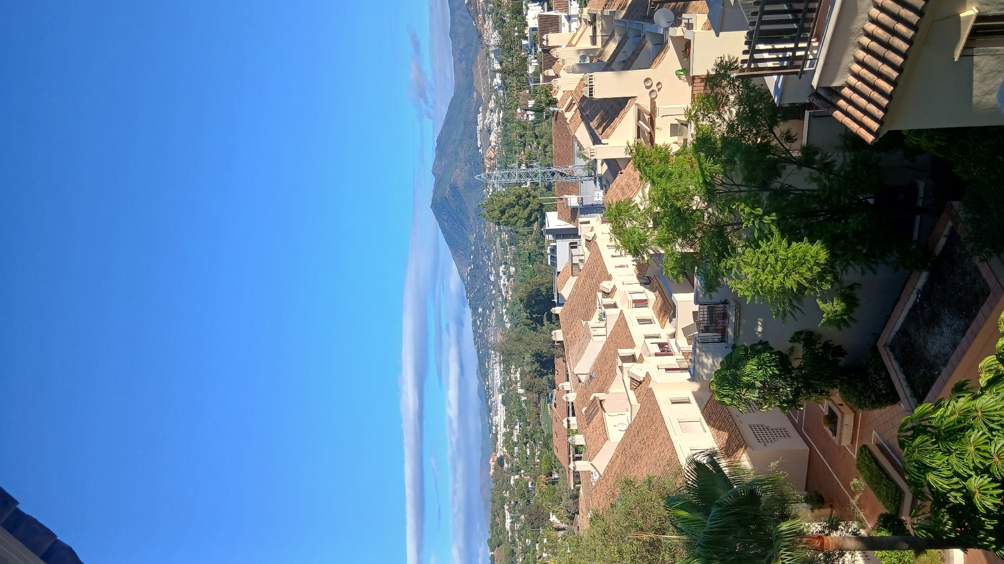 The image is an outdoor, elevated, vertical shot showcasing a cityscape with a mountain range in the background. The foreground is dominated by the terracotta tiled roofs of several residential buildings, typical of a Mediterranean architectural style. These buildings are light-colored, with visible windows and chimneys. Lush green trees are interspersed among the buildings, some featuring broad leaves and others more feathery foliage. A palm tree is visible on the left side of the frame. In the midground, there's a densely populated area with more buildings and trees. Further back, the landscape rises into rolling hills covered in vegetation and scattered white structures, leading to a prominent, conical mountain peak in the distance, its slopes appearing arid. The sky is a clear, bright blue, with a few thin, elongated clouds streaking horizontally across the upper portion of the image. A lattice-like communication tower is visible in the mid-distance, breaking the otherwise organic shapes of the landscape. The lighting suggests it is daytime, likely late morning or early afternoon, given the angle of the sun and the clear sky.