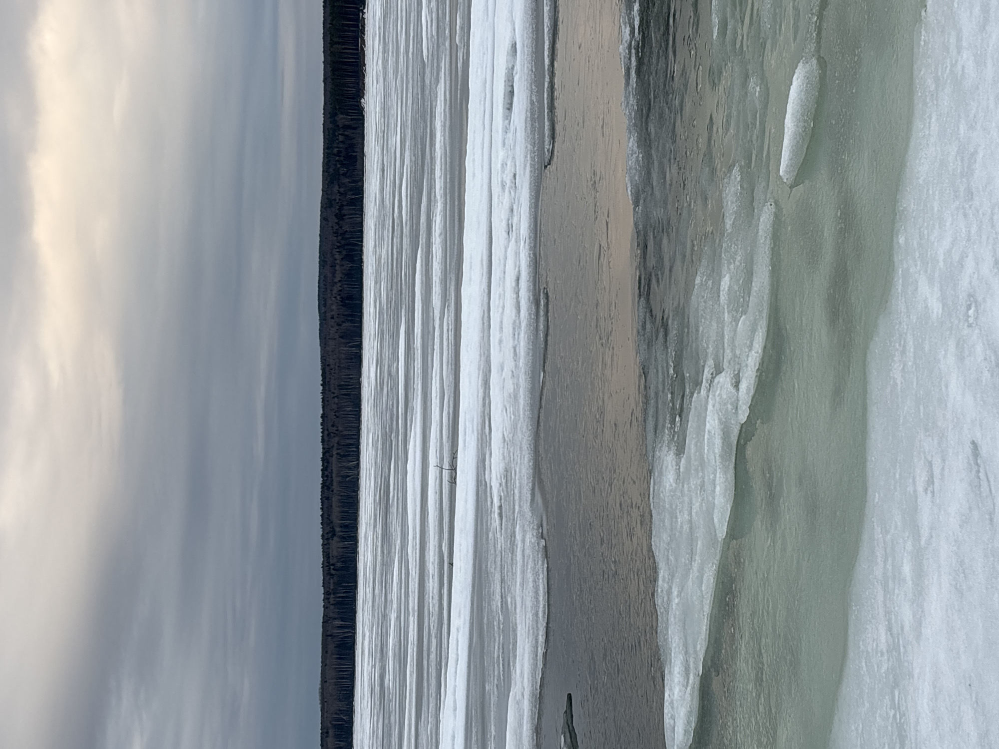 A landscape photograph depicts a frozen body of water, likely a river or lake, with ice formations along the shoreline. The image includes the sky with a cloud cover, and a thin line of trees on the opposite shore. The photo may have been taken in Chaikovsky, Russia.