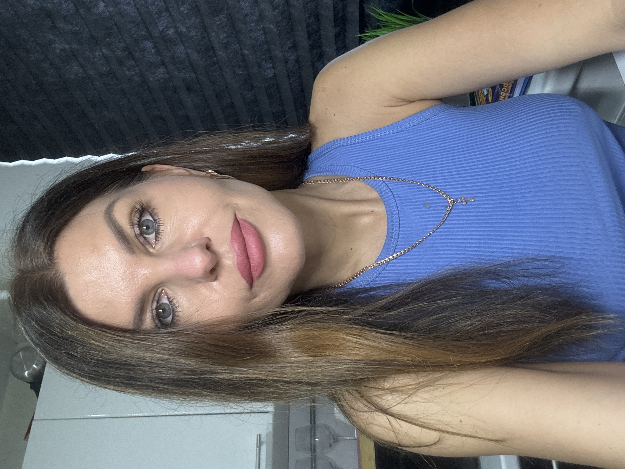 A woman is facing the camera in a close-up shot. She has brown hair with lighter highlights and gray eyes. She wears a blue ribbed tank top and a gold necklace with a cross pendant. Her lips are colored with a pink lipstick. The background includes a dark, vertically textured surface above and a glimpse of a white cabinet and possibly a metallic object below. The location in the city of Chaikovsky, Russia, is not discernible from the image.