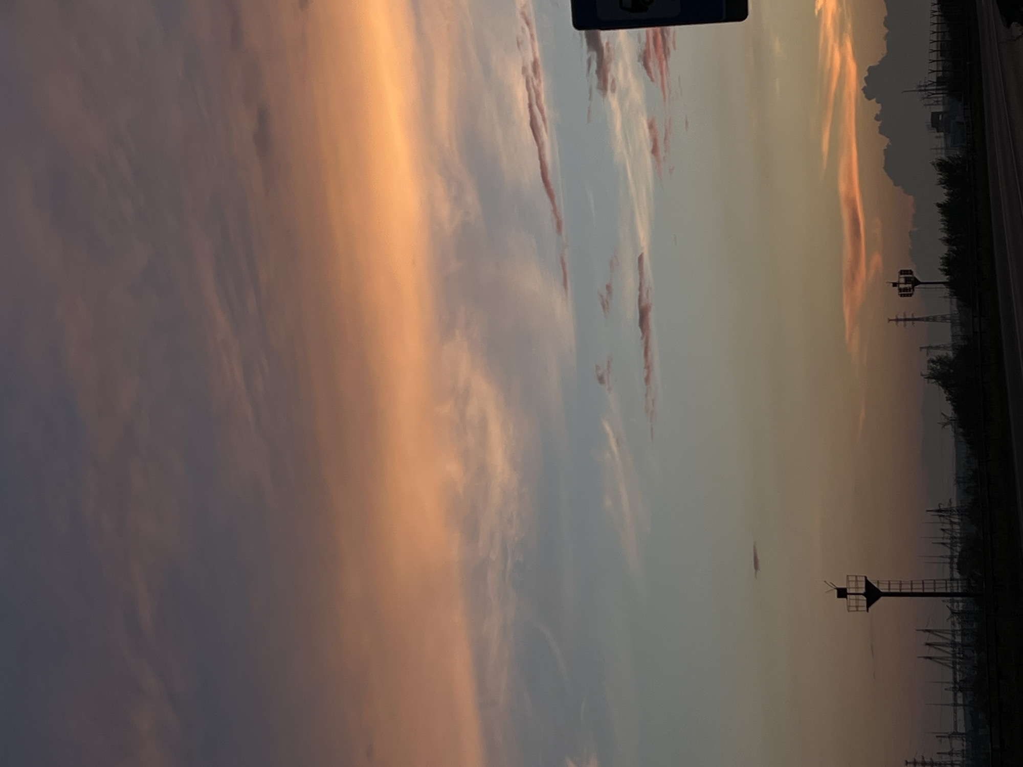 The image captures a sunset over Chaikovsky, Russia, likely near a road or industrial area. The sky is a vibrant display of colors, ranging from deep purple and gray at the top to soft pinks and oranges, transitioning to a pale yellow and gray near the horizon. Wispy clouds are scattered across the sky, some tinged with pink from the setting sun.
In the foreground, silhouetted against the colorful sky, is a dark landscape. A road or paved surface runs horizontally across the bottom of the image. To the left, a tall, thin tower with a ladder and a square platform at the top is visible. Further back and to the left, a series of electrical towers and infrastructure stretch across the horizon, suggesting an industrial zone. In the distance, darker, more defined clouds are present, and the silhouette of a hillside or distant structures can be seen.
To the right of the frame, a dark, rectangular signpost is partially visible, with a circular blue and white emblem at the top. The overall impression is one of a tranquil evening scene, juxtaposing the natural beauty of the sunset with the man-made elements of the urban or industrial landscape. No people or animals are discernible in the image.