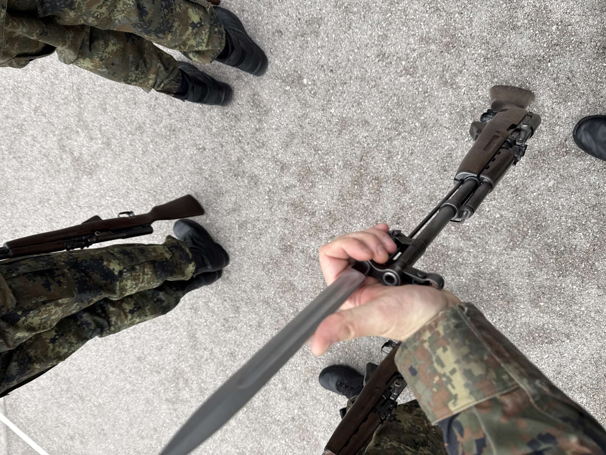 The image depicts three individuals in camouflage uniforms standing on a grey concrete surface, possibly a street or courtyard. Two of the individuals are holding rifles; one holds the rifle, appearing to be inserting a bayonet. The camouflage pattern is multi-toned green, brown, and black. All individuals are wearing black boots. The rifles appear to be of different models, with one having a wooden stock. It is not possible to confirm the location as Sofia, Bulgaria, or provide any more information without further context.