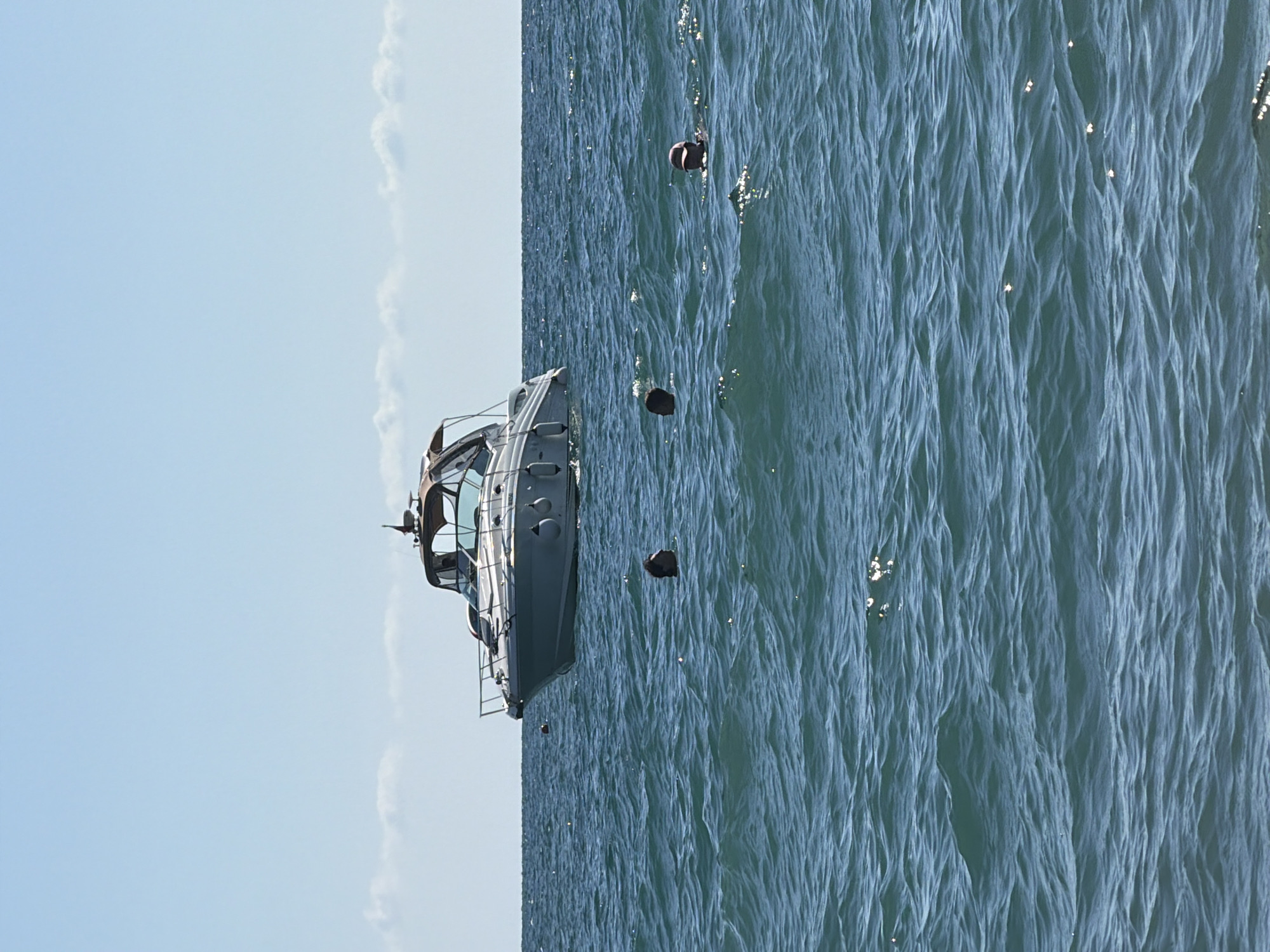 A pleasure boat is anchored in the blue sea off the coast of Sochi, Russia. Several people are swimming in the water near the boat. The sky is clear and blue with a few scattered clouds. The water is choppy with small waves.