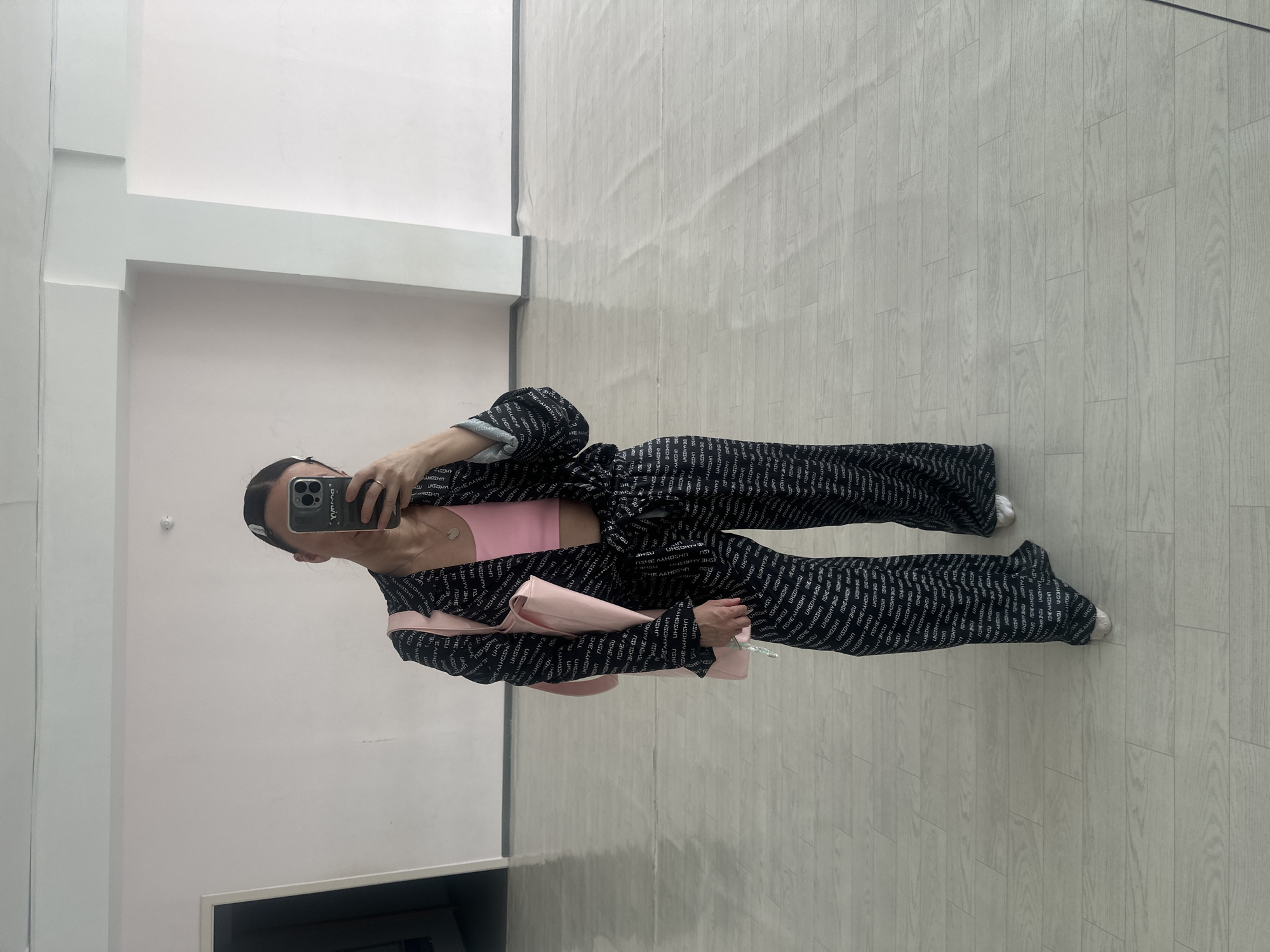 A full-body shot shows a woman holding a cell phone, presumably taking a mirror selfie. She is wearing a black and white patterned pantsuit with a pink crop top underneath. She has a pink purse slung over her shoulder. The setting appears to be an indoor room with a light-colored wooden floor and white walls. The location cannot be determined from the image, and there is no information to suggest it is in Votkinsk, Russia.