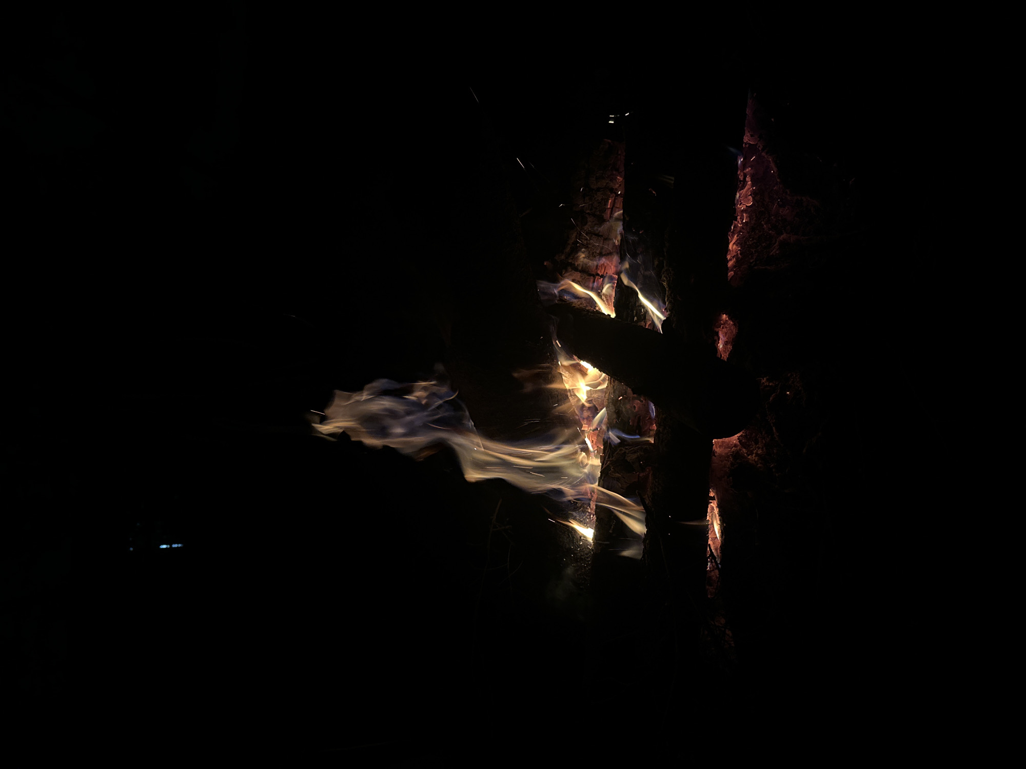 The image shows a campfire at night. It appears to be constructed of logs arranged in a vertical stack, with flames and embers visible. The environment is dark, indicating an outdoor location. While I can interpret visual information, I don't have the capacity to pinpoint the location as Юзуфоўскі сельскі Савет, Belarus. There are no visible people in the image.