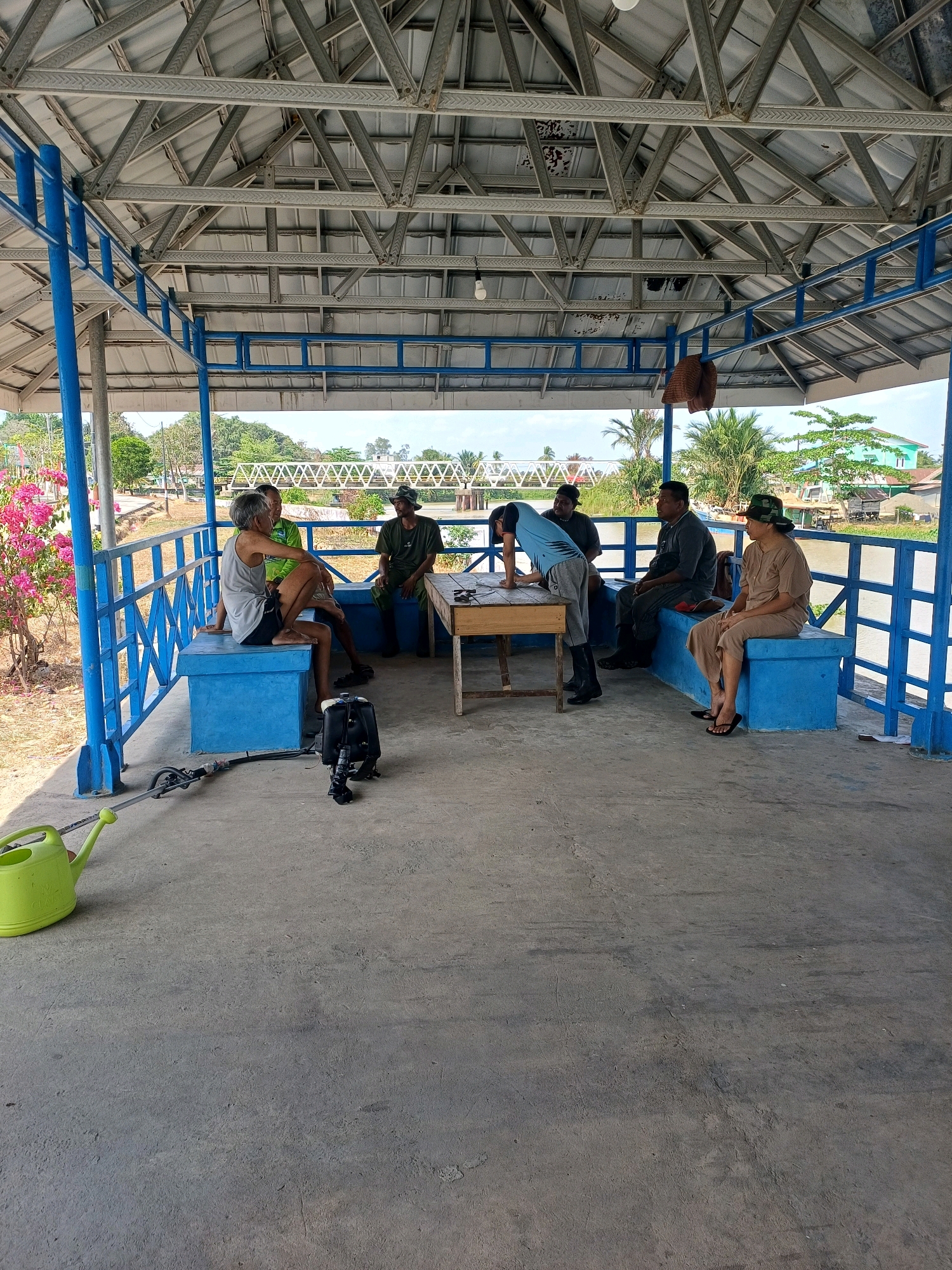 In Pasir Belengkong, Indonesia, a group of seven individuals are gathered under a covered, open-air structure with a metal roof supported by blue metal beams. The structure appears to be situated near a body of water, with a bridge visible in the background, along with trees and buildings. Five men and two women are present.
On the left side of the structure, two men are seated on a blue bench. One man is wearing a grey tank top and shorts, while the other is in a green short-sleeved shirt. A backpack and what appears to be a grass trimmer are placed on the concrete floor near them.
In the center, a wooden table is positioned. Three men are gathered around it. One man, wearing a blue shirt and grey pants, is bent over the table, seemingly examining something. Another man, dressed in a dark shirt, stands behind him. A third man, in a grey long-sleeved shirt, is seated on a blue bench to the right of the table.
On the far right, a woman is seated on a blue bench, wearing a tan outfit and a hat. She appears to be looking towards the table. A fourth man, wearing a green uniform and a hat, is seated on the same bench as the woman.
The overall scene depicts a casual gathering, possibly for discussion or a break, in an outdoor setting within the vicinity of Pasir Belengkong.