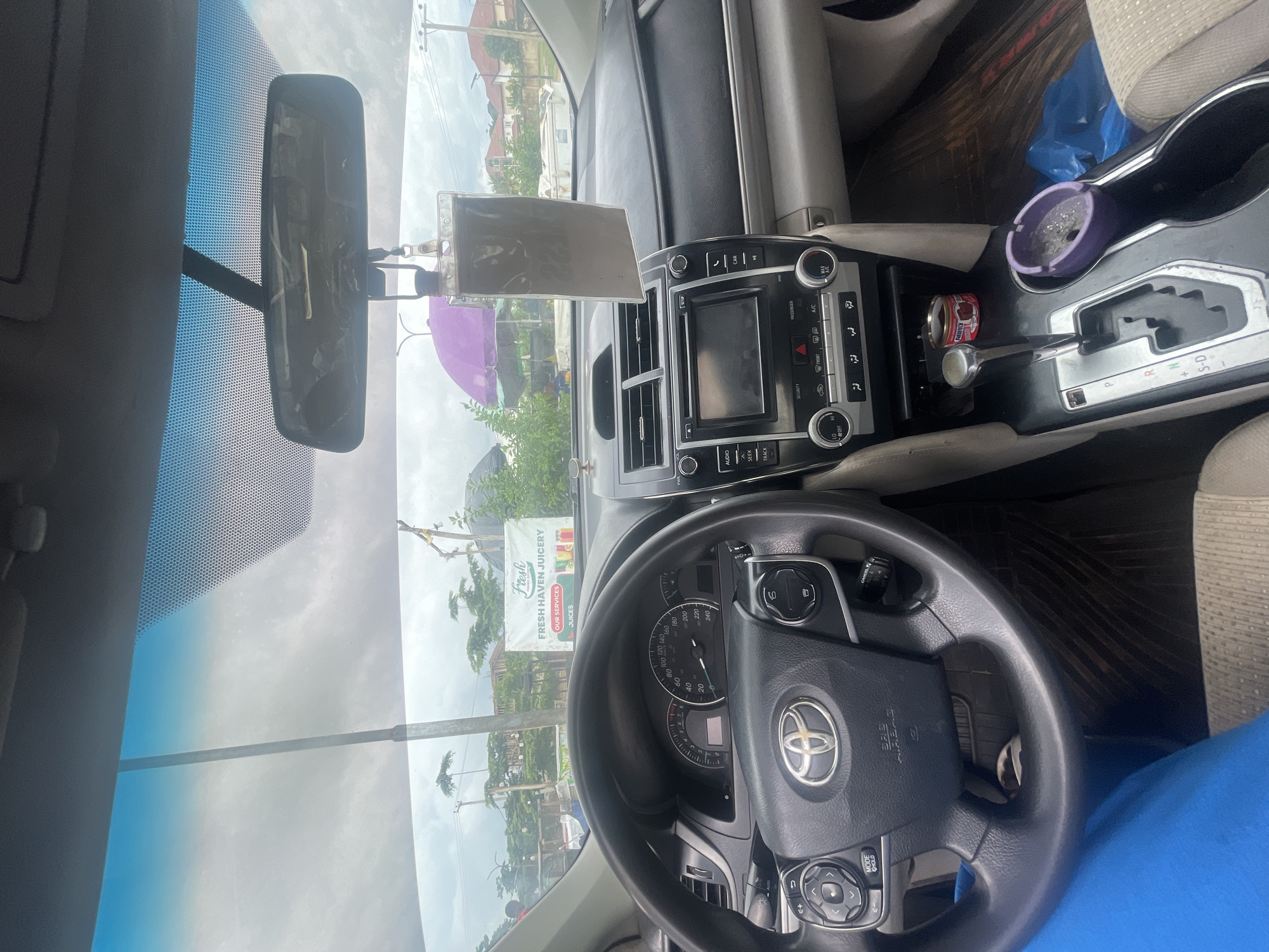 The image shows the interior of a car, viewed from the driver's perspective. The car is a Toyota Camry, identifiable by the emblem on the steering wheel. The dashboard features a multimedia system, climate controls, and a speedometer. A car air freshener hangs from the rearview mirror.
Outside the car, visible through the windshield, is a street scene in Galadimawa Village, Nigeria. A banner for "Fresh Haven Juicery" is displayed, along with an umbrella and some trees. The sky is overcast. There are no people clearly visible within the car's interior.