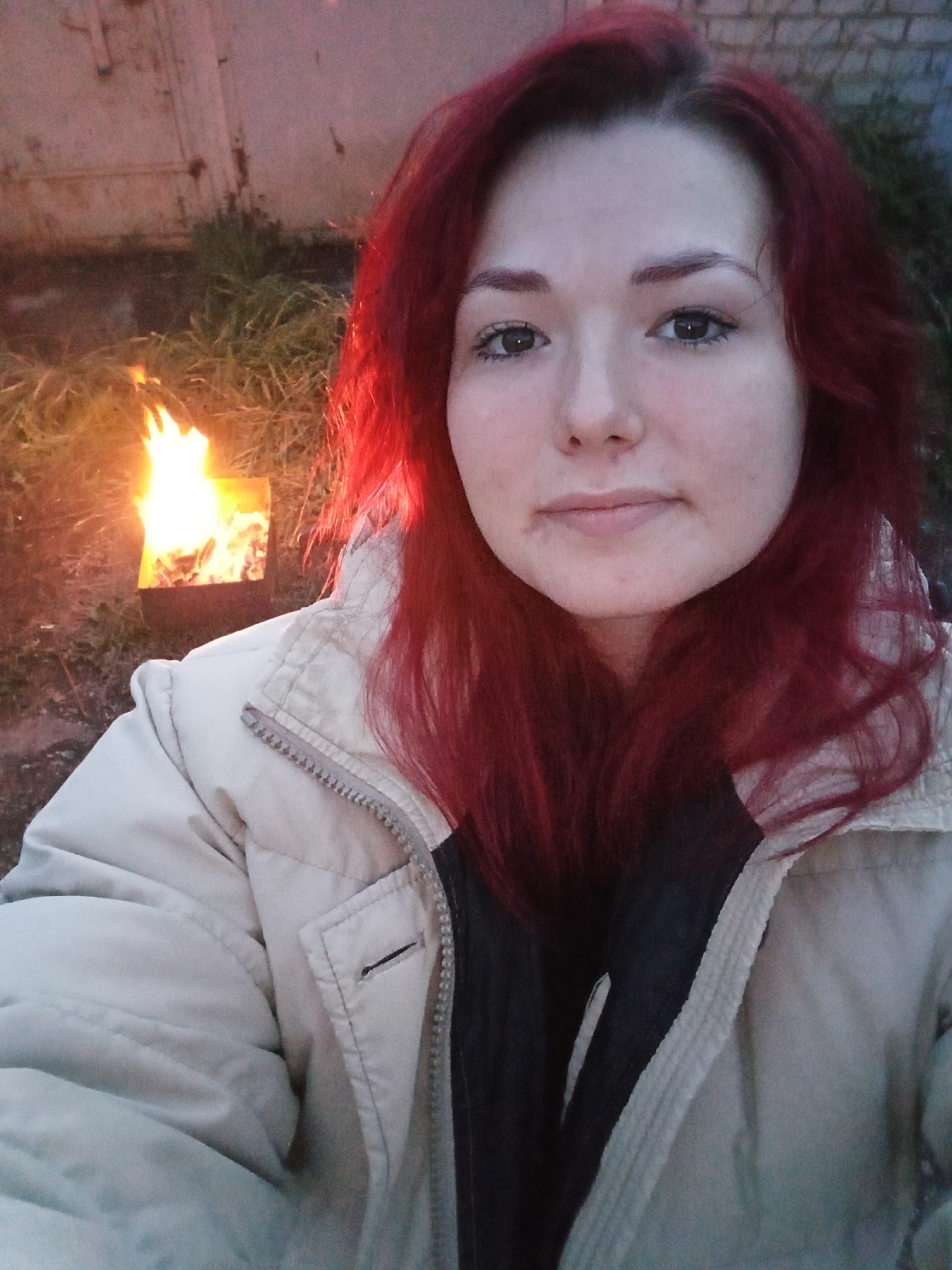 In this self-portrait, a young woman with vibrant red hair and light skin is positioned in the foreground, looking directly at the camera. She is wearing a light-colored, possibly beige or cream, quilted jacket with a gray zipper. Her hair cascades around her shoulders, framing her face. To her left, and slightly behind her, a small metal brazier contains a bright, roaring fire. The background consists of dry, light-colored grass or straw and a rough, textured surface that appears to be a brick wall. The scene suggests an outdoor setting, possibly a backyard or an area with a makeshift fire pit, in the vicinity of a brick structure. The overall lighting indicates it is either dusk or dawn, with the fire providing a significant light source.