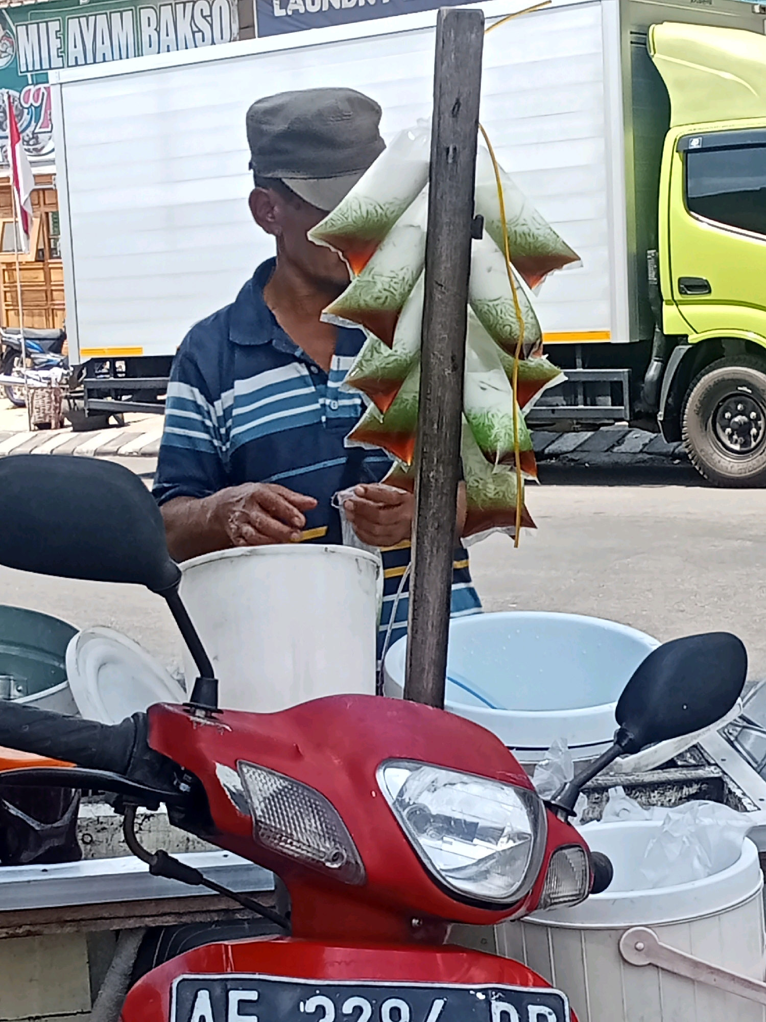 A street vendor in Sukowinangun, Indonesia, is seen in front of a white delivery truck and a bright yellow truck. The vendor, wearing a blue and white striped shirt and a grey cap, is holding plastic bags filled with a beverage. These bags are hanging from a wooden pole. In the foreground, a red scooter is parked, with its handlebars and headlight visible. Various buckets and containers are also present in the scene, suggesting the vendor is selling drinks or food. The backdrop is a typical street environment with a paved road and sidewalk.