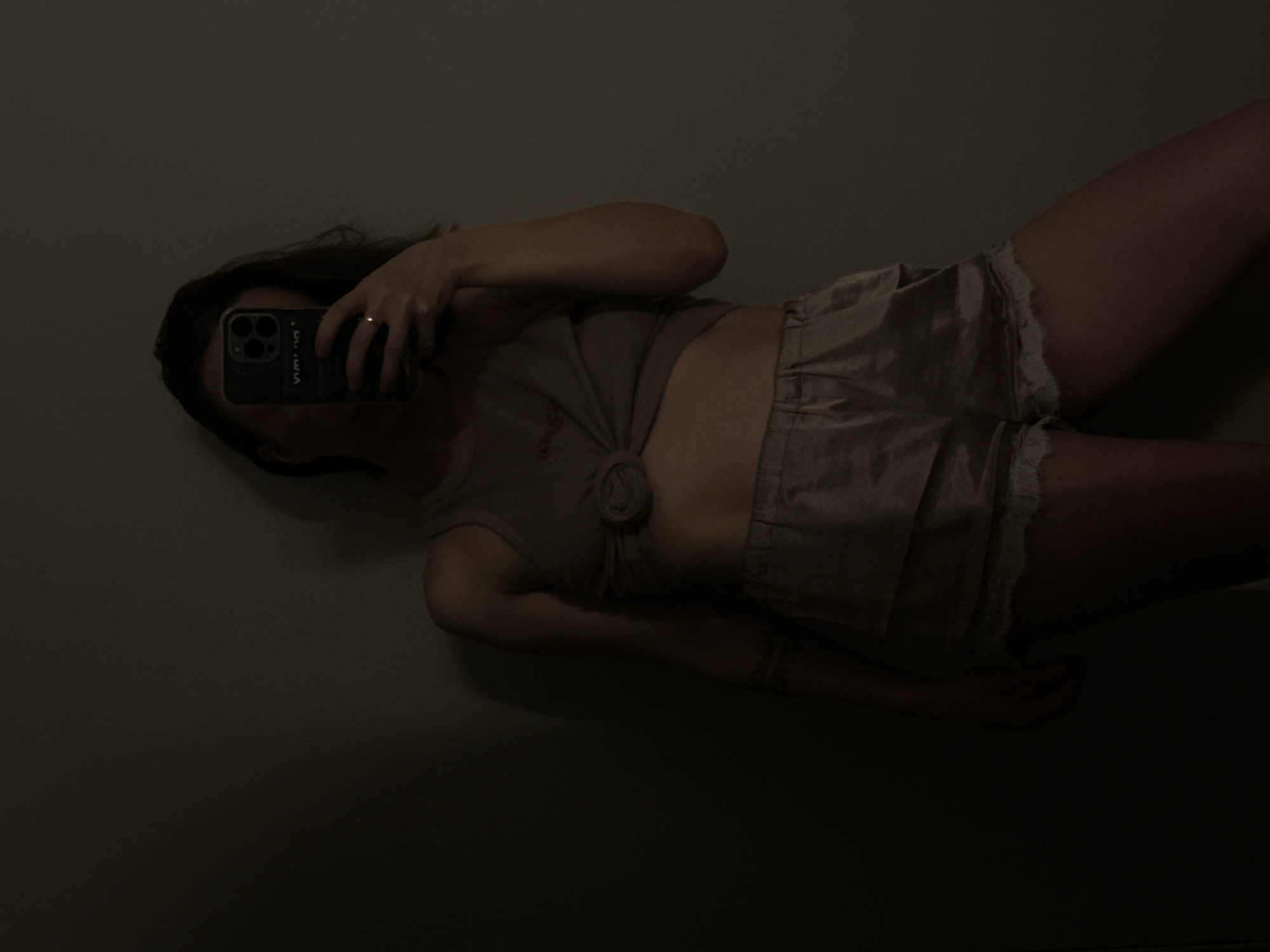 A person is holding a smartphone with multiple lenses to take a selfie. The person is wearing a top that is tied at the stomach and shorts with lace trim. The environment appears to be indoors and dimly lit. There is no information to suggest the location is Votkinsk, Russia.