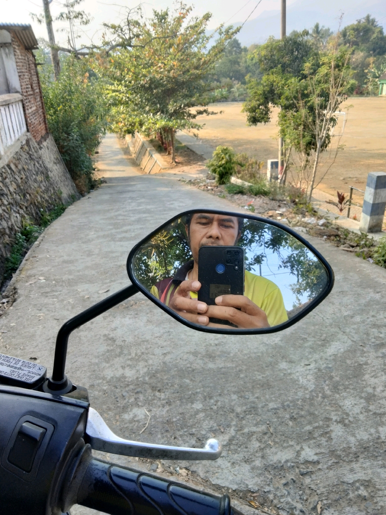 A mirror-image selfie is taken by a person sitting on a motorcycle in Sawahan, Indonesia. The person, wearing a yellow t-shirt, holds a black smartphone to capture their reflection. The motorcycle's handlebar and brake lever are visible in the foreground. The background shows a paved road winding uphill, bordered by a brick wall and lush green trees. In the distance, a field and more trees are visible under a bright sky. The scene appears to be in a rural or semi-rural area.