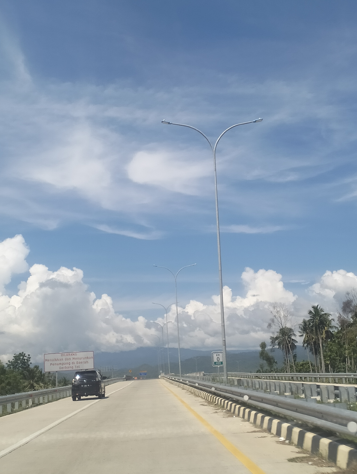 The image captures a bright, daytime scene on a highway bridge in Aron Bunoet, Indonesia. The sky is a vibrant blue, dotted with fluffy white clouds. Several modern streetlights with curved poles are visible, lining the highway. On the right side of the road, there's a metal guardrail with alternating black and white striped concrete barriers. Beyond the guardrail, a line of palm trees and other foliage marks the edge of the landscape.
In the foreground, a black SUV is driving on the highway. To the left of the road, a white sign with red text is posted. The text on the sign appears to be a warning, with the word "DILARANG" (prohibited) visible at the top. Further down the highway, a green sign indicating "KM 0" is also visible. The road surface is asphalt, with clear lane markings. The overall impression is one of a well-maintained highway with a tropical landscape in the background.
