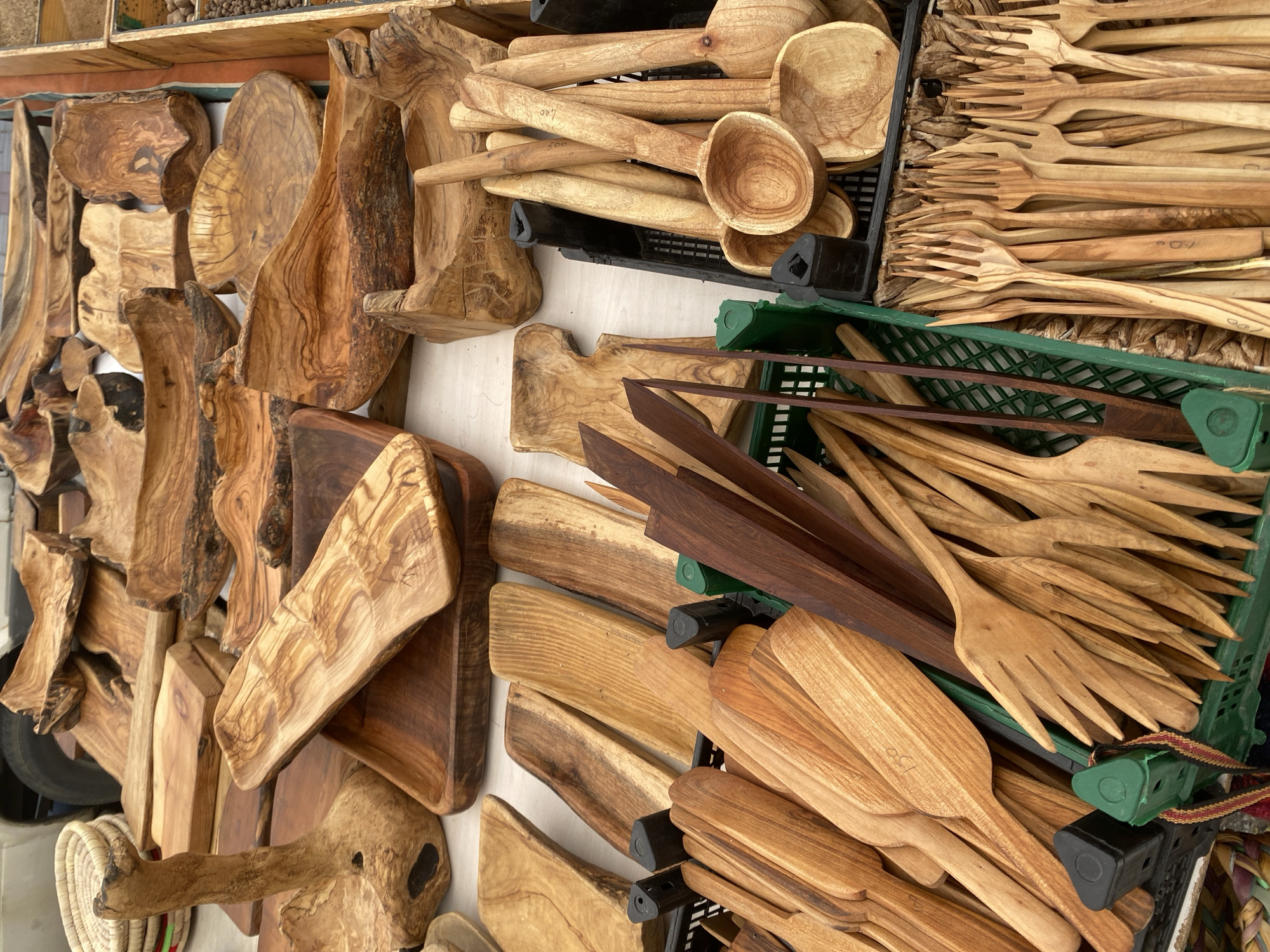 A high-angle, eye-level shot shows an outdoor display of handcrafted wooden kitchen utensils for sale, likely at a market or shop, possibly in Alanya, Turkey. The items, resting on a white surface, include:
* Wooden cutting boards in various shapes and sizes, some with live-edge detailing.
* Assorted wooden spoons, some stacked in a black plastic crate.
* Wooden forks, also in a black plastic crate.
* A collection of spatulas, possibly olive wood, nestled in a green plastic crate.
The lighting is natural and bright, casting shadows and highlighting the wood grain. The products are the primary focus.