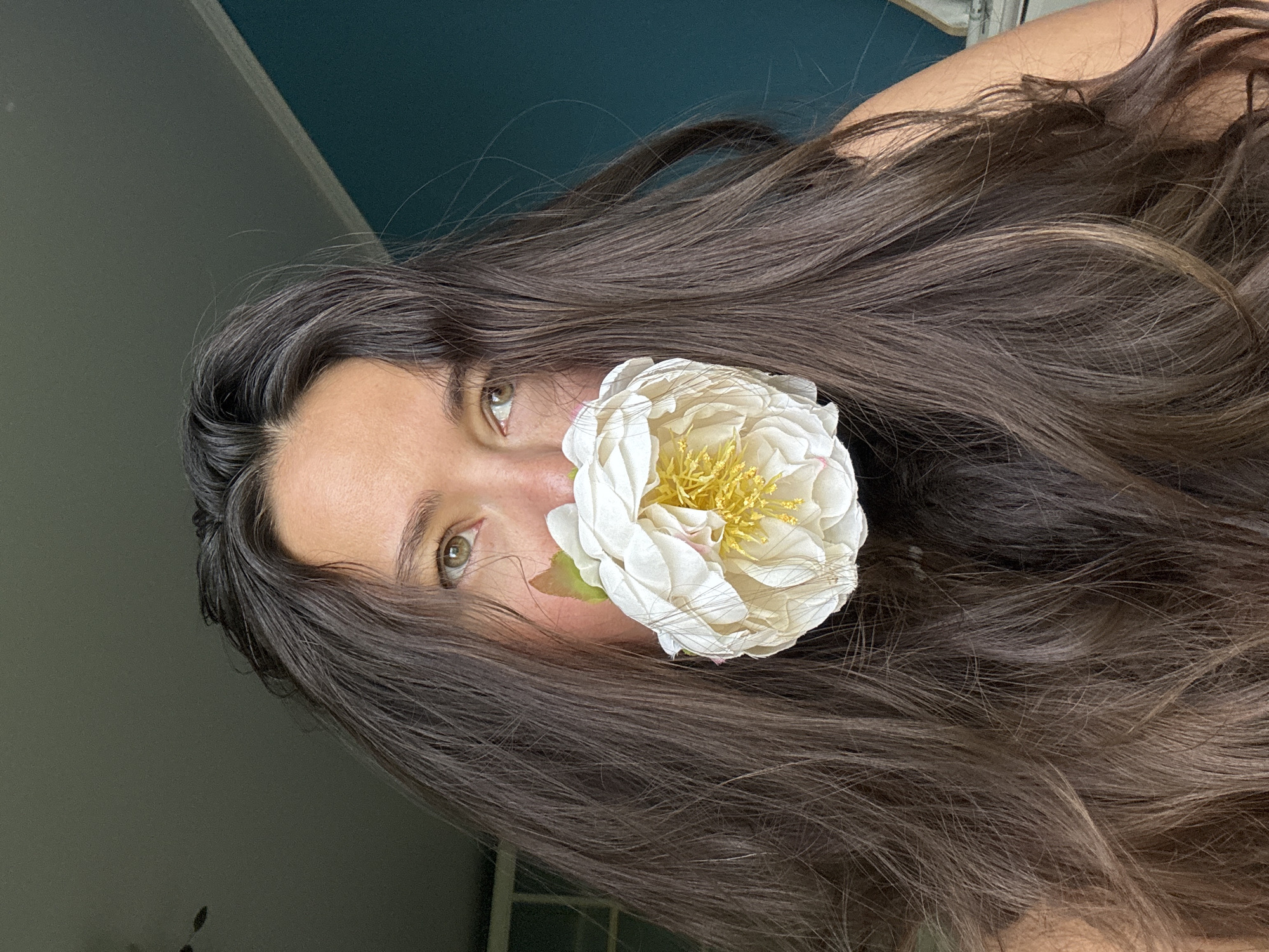 A headshot of a woman with long, dark brown hair. Her hair falls around her face and over her shoulder. A white and yellow flower is positioned near her face. The woman has light skin and light-colored eyes. The background shows a light gray wall and a section of a dark blue wall. The image is rotated counter-clockwise. The location is not specified.