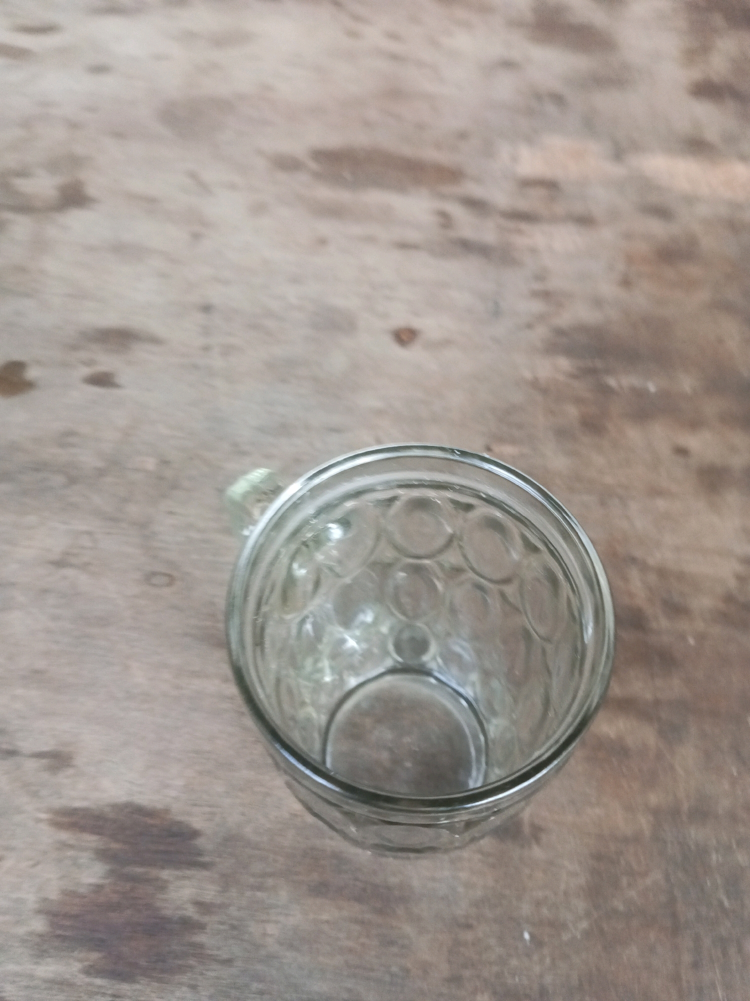 An empty, clear glass mug with a patterned interior rests on a wooden surface. The mug has a handle on the left side. The wooden surface appears to be a table with visible grain and some water stains. The image was taken outdoors, likely in the city of South Tangerang, Indonesia, judging by the context provided. There are no people or other objects in the frame.