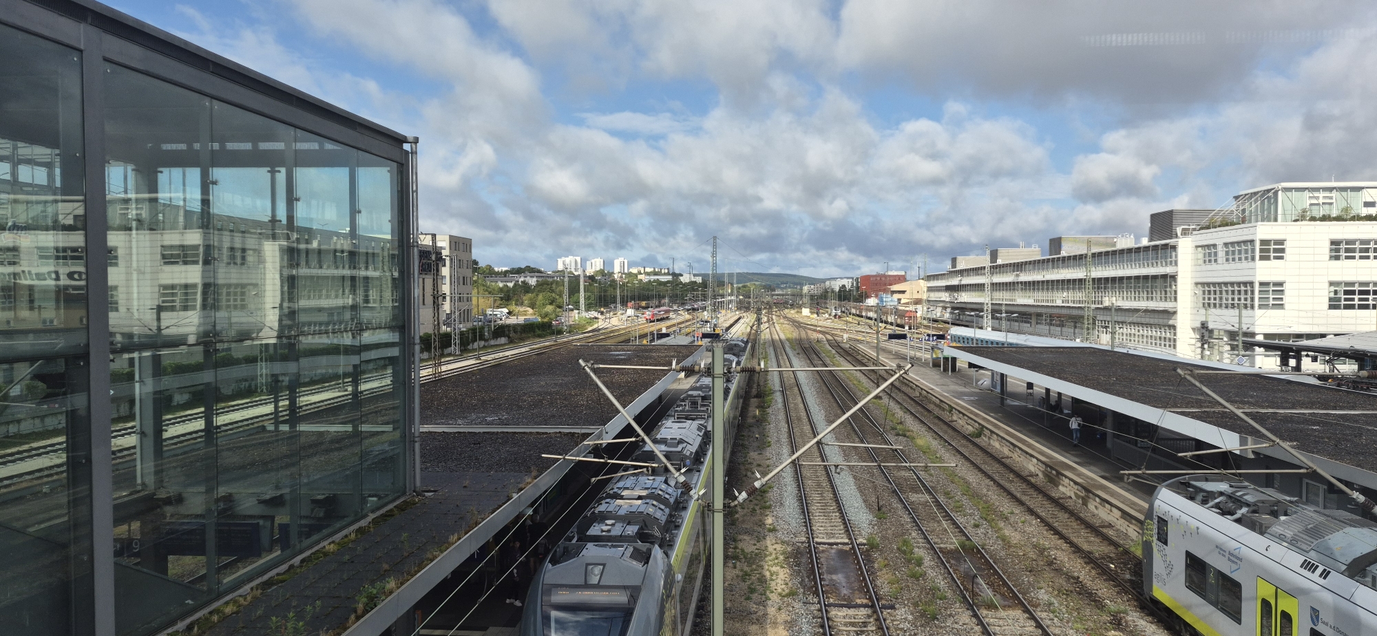 The image depicts a view of the Regensburg Hauptbahnhof (main train station) in Regensburg, Germany. In the foreground on the left is a modern building with a glass facade, reflecting the sky and surrounding structures. To the right of this building are multiple train tracks, with a modern gray and yellow train occupying one of the platforms. Another train, primarily gray, is visible further down the tracks. In the background, a complex network of railway lines converges, and beyond that, the cityscape of Regensburg is visible, characterized by apartment buildings and other urban structures against a backdrop of hills under a cloudy sky. The scene captures the activity and infrastructure of a busy train station.