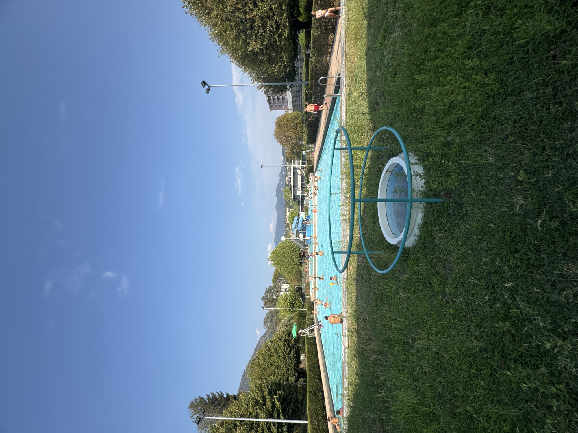 The image depicts an outdoor swimming pool complex on a sunny day, with a clear blue sky and scattered clouds. The pool is filled with people, many of whom are swimming or wading in the water. In the foreground, a grassy area is visible with a circular concrete structure surrounded by a blue metal railing, possibly a decorative element or part of the pool's filtration system.
To the left of the pool, lush green trees and manicured hedges line the perimeter. Beyond the pool, a lifeguard station with a green umbrella and a tall white chair stands near the water's edge. In the background, buildings are visible, suggesting an urban or suburban setting. The presence of a waterslide indicates the facility is likely a public leisure center. A mountain range is visible in the distance, partially obscured by trees. The overall scene conveys a vibrant and active atmosphere, typical of a summer day at a swimming pool. The location appears to be the "Piscina Comunale di Chiasso" in Chiasso, Switzerland, given the architectural style of the background buildings and the surrounding landscape.