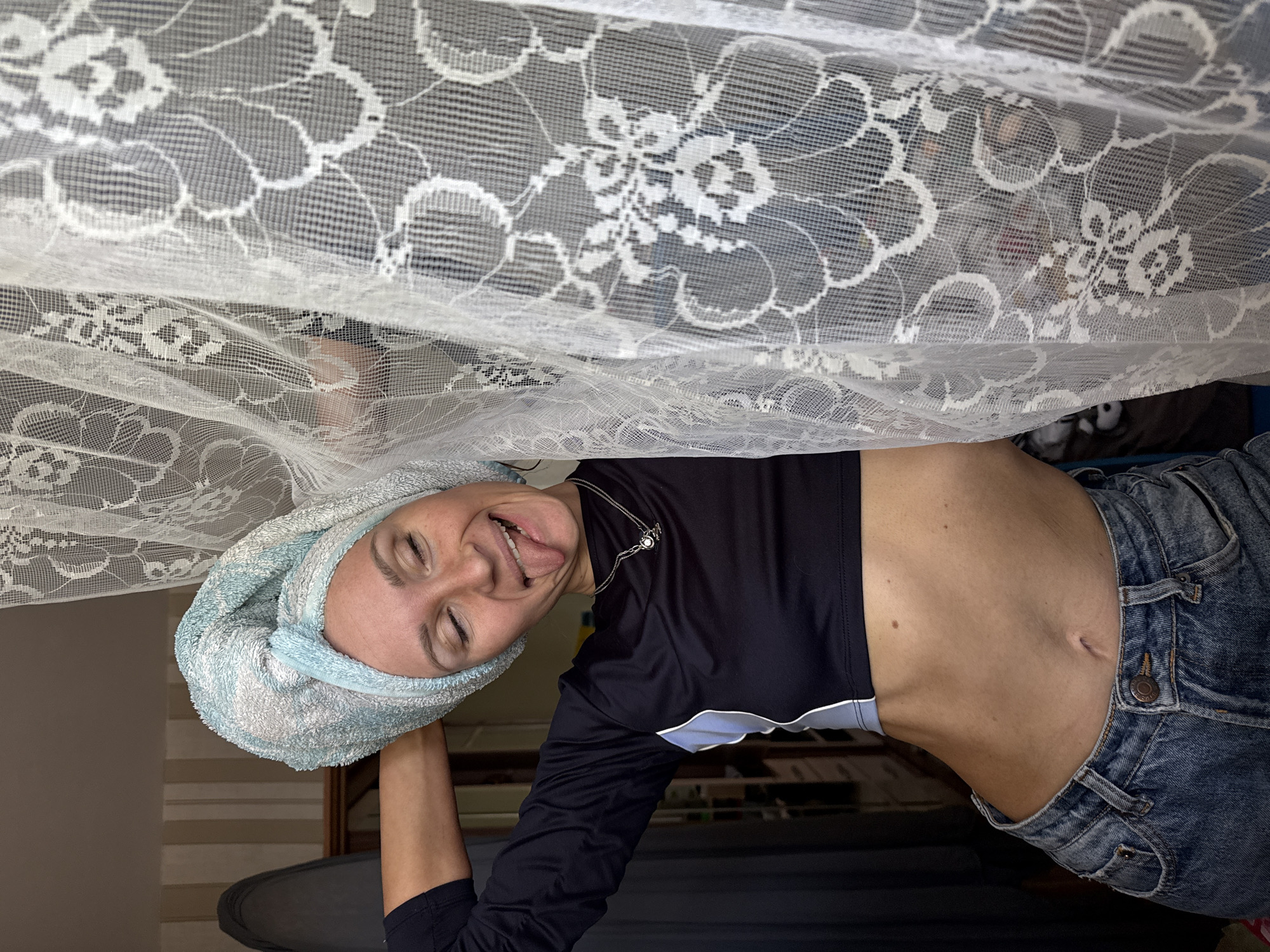 A woman with a blue towel wrapped around her head is looking at the camera, sticking her tongue out. She is wearing a dark blue, long-sleeved cropped shirt, blue jeans, and a silver necklace with a circular pendant. She appears to be standing behind a white lace curtain. The environment suggests an indoor location in Chaikovsky, Russia.