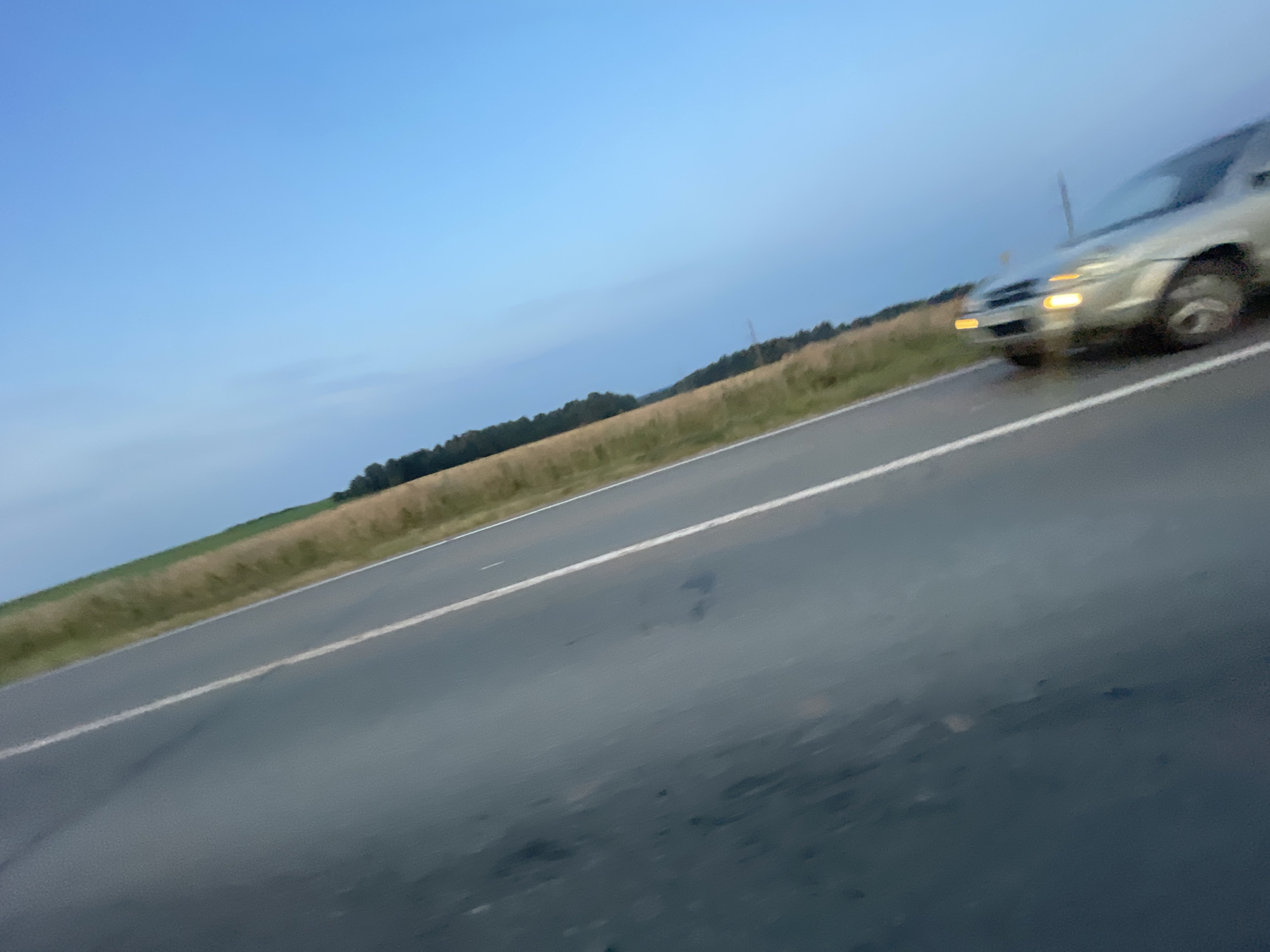 The image shows a desolate stretch of wet asphalt road with a white line dividing it. To the right of the road, there is a grassy verge and then a treeline. The sky is a clear, bright blue, suggesting it is daytime, possibly late afternoon or early morning. In the lower right portion of the image, a silver car is visible, appearing to be stationary. The car's headlights are illuminated, and its reflection is visible on the wet road surface. The image appears to be taken from a moving vehicle or an elevated perspective, as the road is angled. There are no people visible in the image. The location is identified as Ol'khovo, Russia.