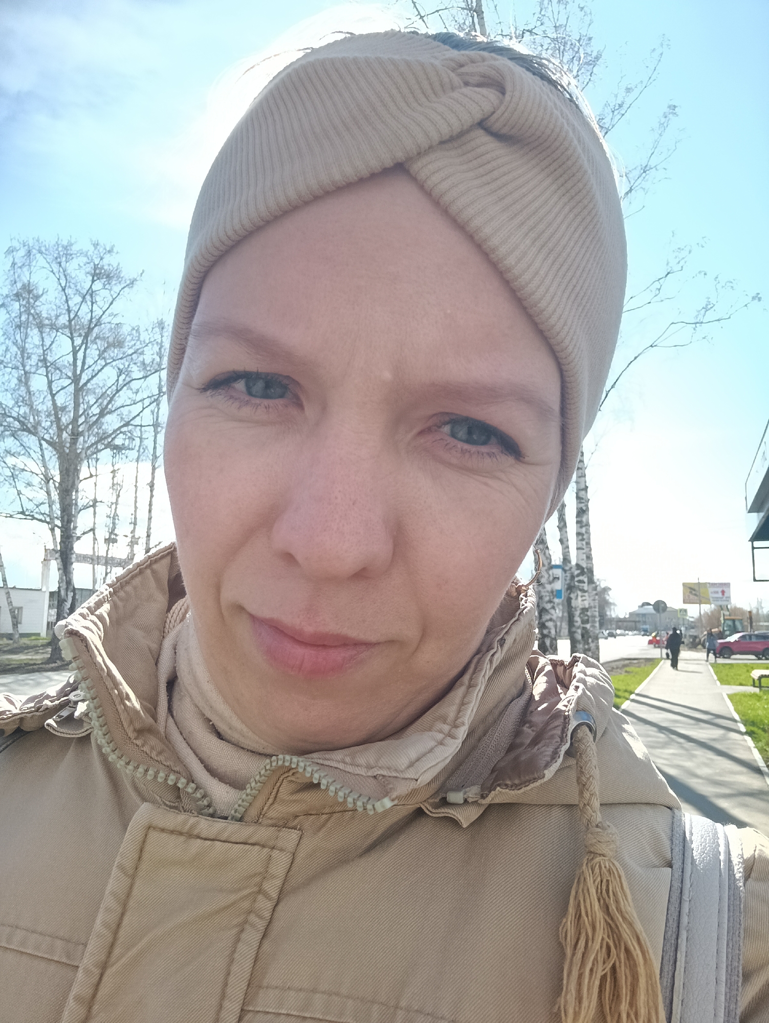 The image is a selfie of an adult woman taken outdoors on a clear, sunny day in Chaikovsky, Russia. The woman, who appears to be light-skinned, is positioned centrally in the frame, looking directly at the camera with a calm, slightly thoughtful expression. She has light blue-grey eyes.
She is wearing a light beige or tan ribbed headband, styled with a decorative knot on top. Her attire includes a light brown or tan zippered jacket, beneath which a matching high collar or scarf is visible. A braided tassel, likely from a hood drawstring, hangs over her right shoulder, and part of a white and grey striped bag is visible on her right side.
The background shows an urban or suburban street scene under a bright blue sky. On the left, several bare-branched trees, possibly birches, line what appears to be a road or open area. Further back, buildings are visible. To the right of the woman, a paved sidewalk stretches into the distance, flanked by a strip of green grass. Several pedestrians can be seen walking along the sidewalk, and vehicles are present on the road. A small yellow vehicle, possibly construction equipment, is partially visible further down the street. The bright sunlight suggests midday and the bare trees indicate either early spring or late autumn. No clearly readable text is discernible on any signs or buildings in the background.