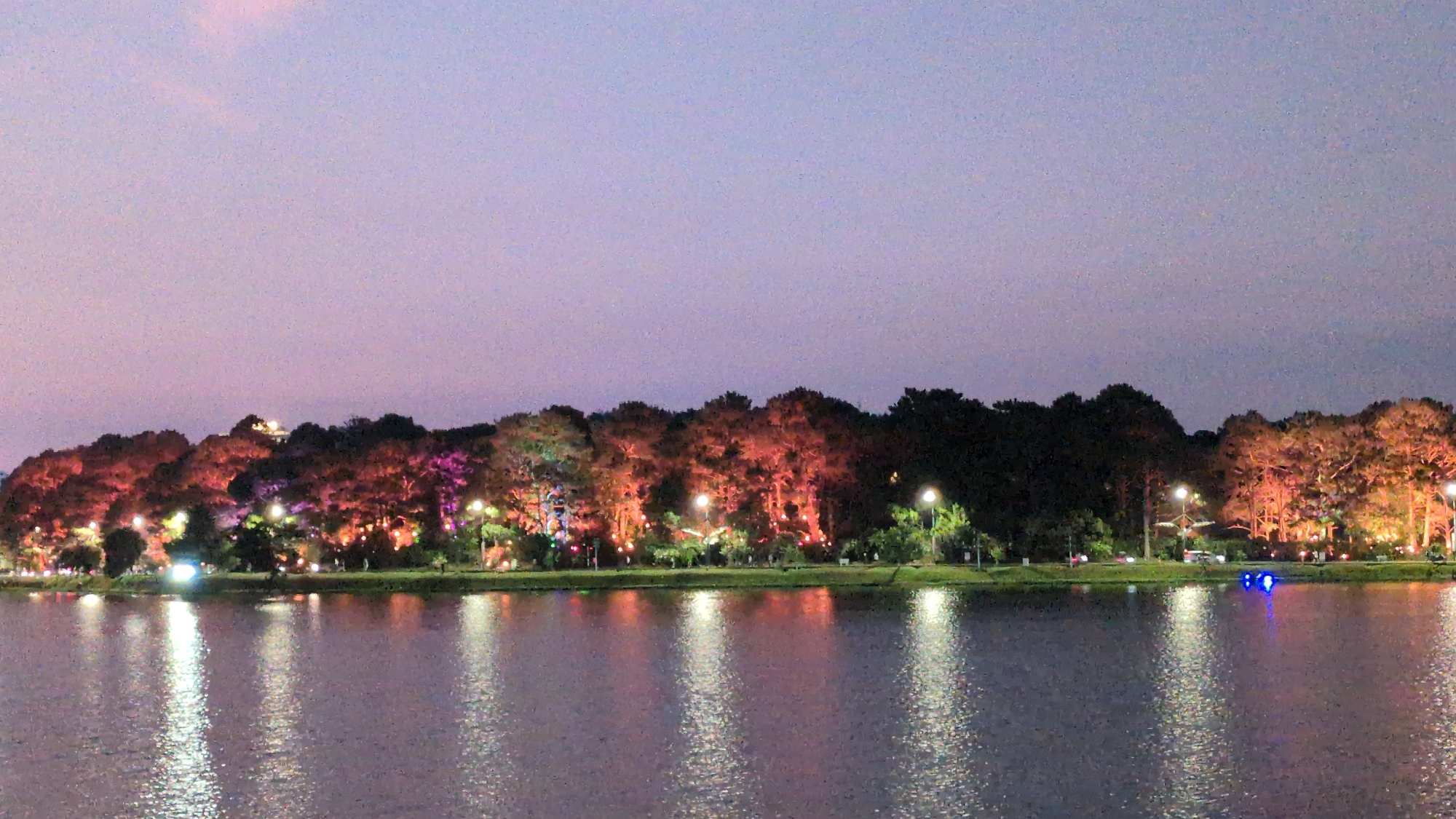 The image depicts a body of water reflecting lights from the opposite shore. The sky transitions from a light purple-pink hue near the top to a slightly darker shade lower down. The shoreline is lined with trees, illuminated with a variety of colored lights, including red, orange, pink, and white. Streetlights are also visible along the shoreline. The lights are reflected in vertical streaks on the water's surface. The location is likely Ho Xuan Huong Lake in Da Lat, Vietnam, judging from the distinctive appearance of the landscape lighting.