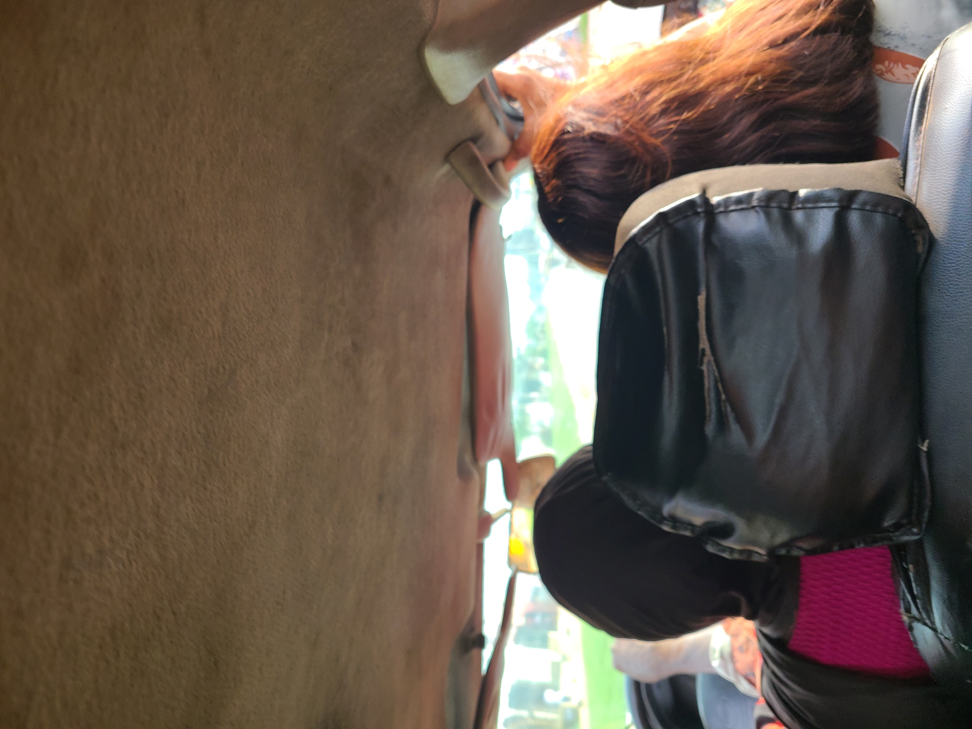 The image, taken from inside a vehicle, shows the backs of two individuals seated in the rear. The person on the left wears a black headscarf and a bright pink top. The person on the right has long, reddish-brown hair. The interior of the vehicle, including the headrests and ceiling, is visible. The exterior view through the windshield is blurred but suggests a daytime setting with greenery. The context of Karu, Nigeria, indicates this scene is likely within that urban or peri-urban area.