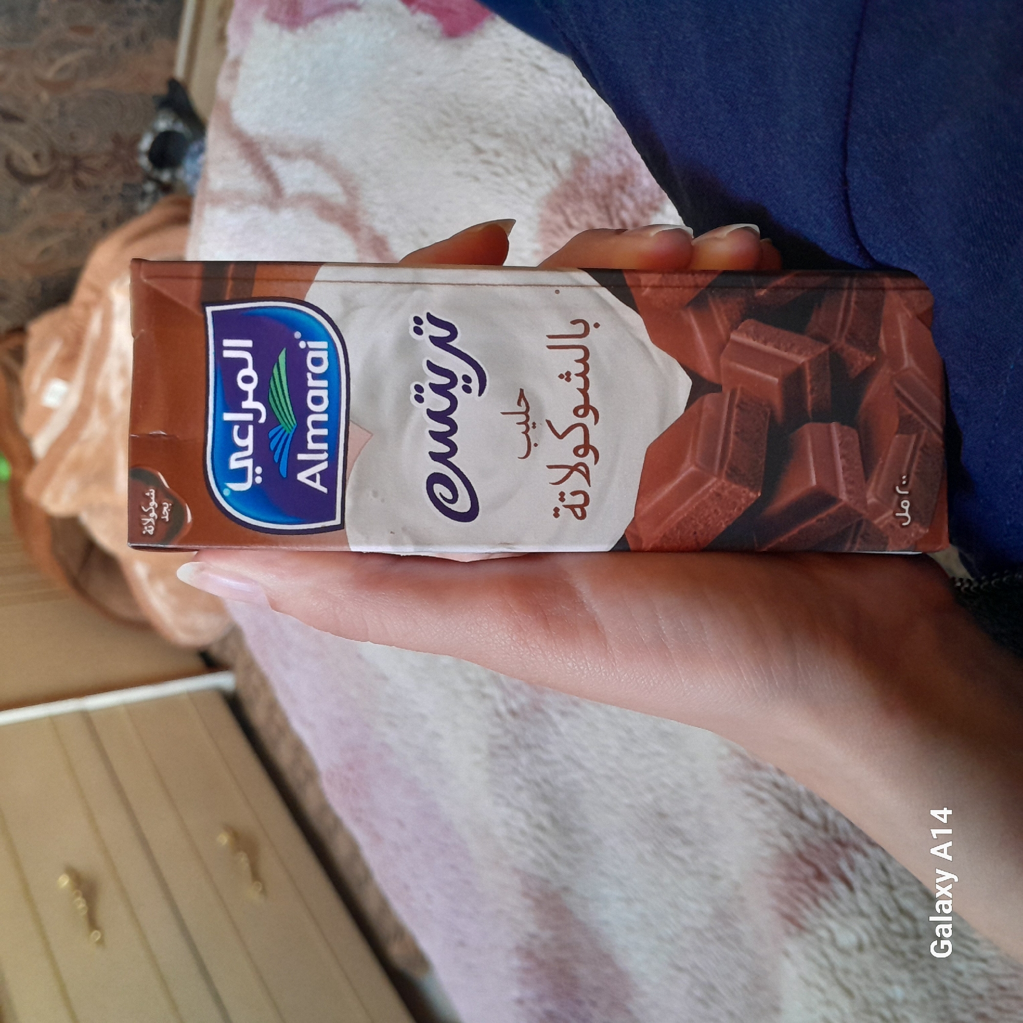 A person holds a carton of Almarai chocolate milk. The carton is brown and white, with a picture of chocolate on one side. The person's hand is visible, with fair skin and long nails. The background is blurry and out of focus, but appears to be a bed with blankets and furniture. The location is unknown.