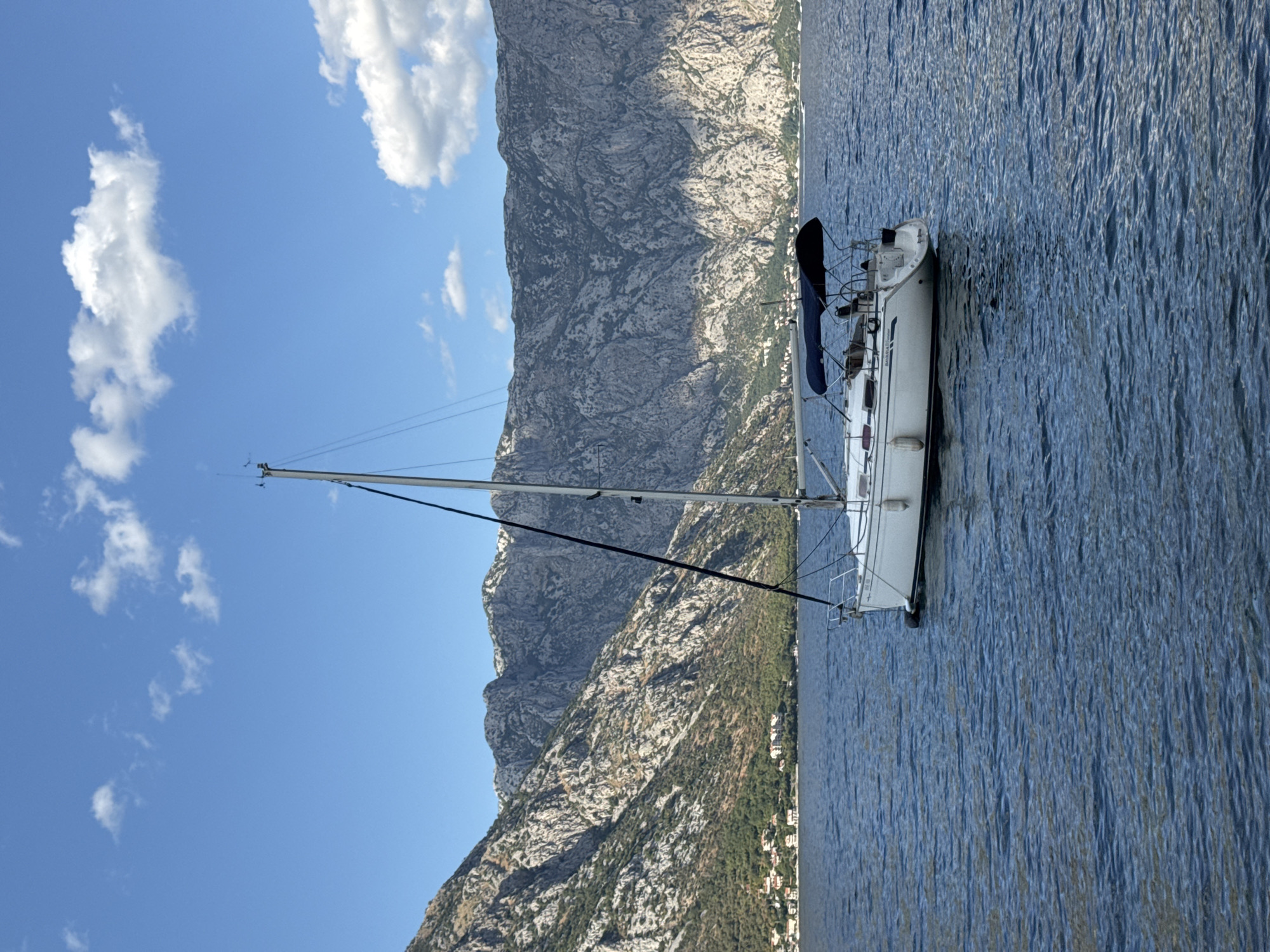 A white sailboat is anchored in the clear blue waters of the Bay of Kotor, near the town of Prčanj, Montenegro. The boat has a tall mast and a dark blue canopy covering the cockpit. In the background, steep, rugged mountains rise dramatically from the coastline. Scattered along the shoreline are small houses with red roofs, characteristic of the region. The sky is a vibrant blue with a few scattered white clouds, indicating a sunny day. The water is calm with gentle ripples. No people are clearly visible on the boat.
