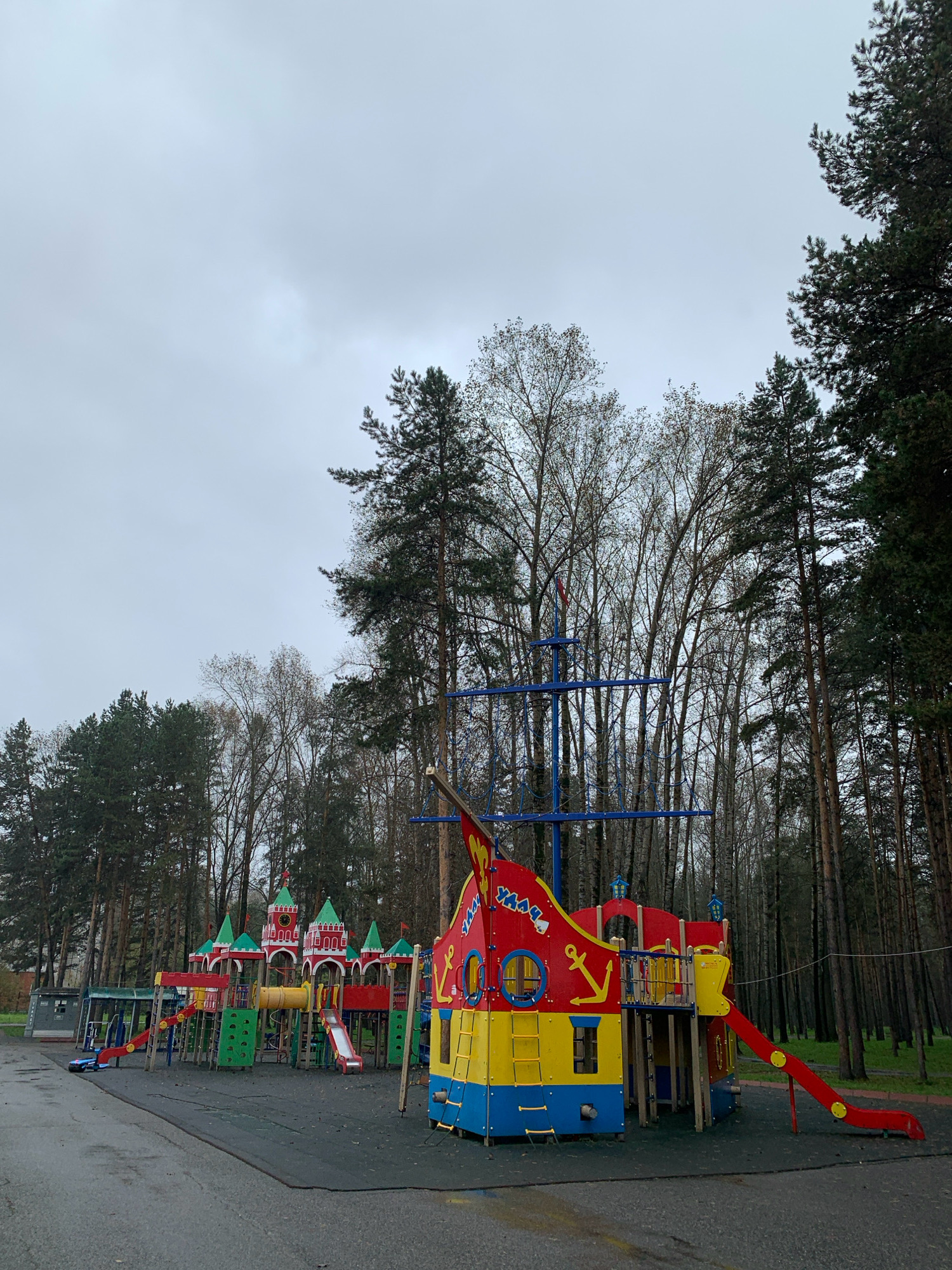 The image depicts an outdoor playground in Prokopyevsk, Russia, on a cloudy day. The playground features a large, colorful ship-themed play structure with red and yellow sides, a blue mast, and an anchor emblem. Adjacent to it is a castle-themed play structure with red walls, green roofs, and a clock face. Both structures are made of plastic and metal, with various slides, ladders, and tunnels. The playground is situated on a dark, rubberized surface, likely for safety. Tall pine trees form a dense forest backdrop behind the playground, with some deciduous trees also visible. The ground is wet, suggesting recent rain. There are no people visible in the image.