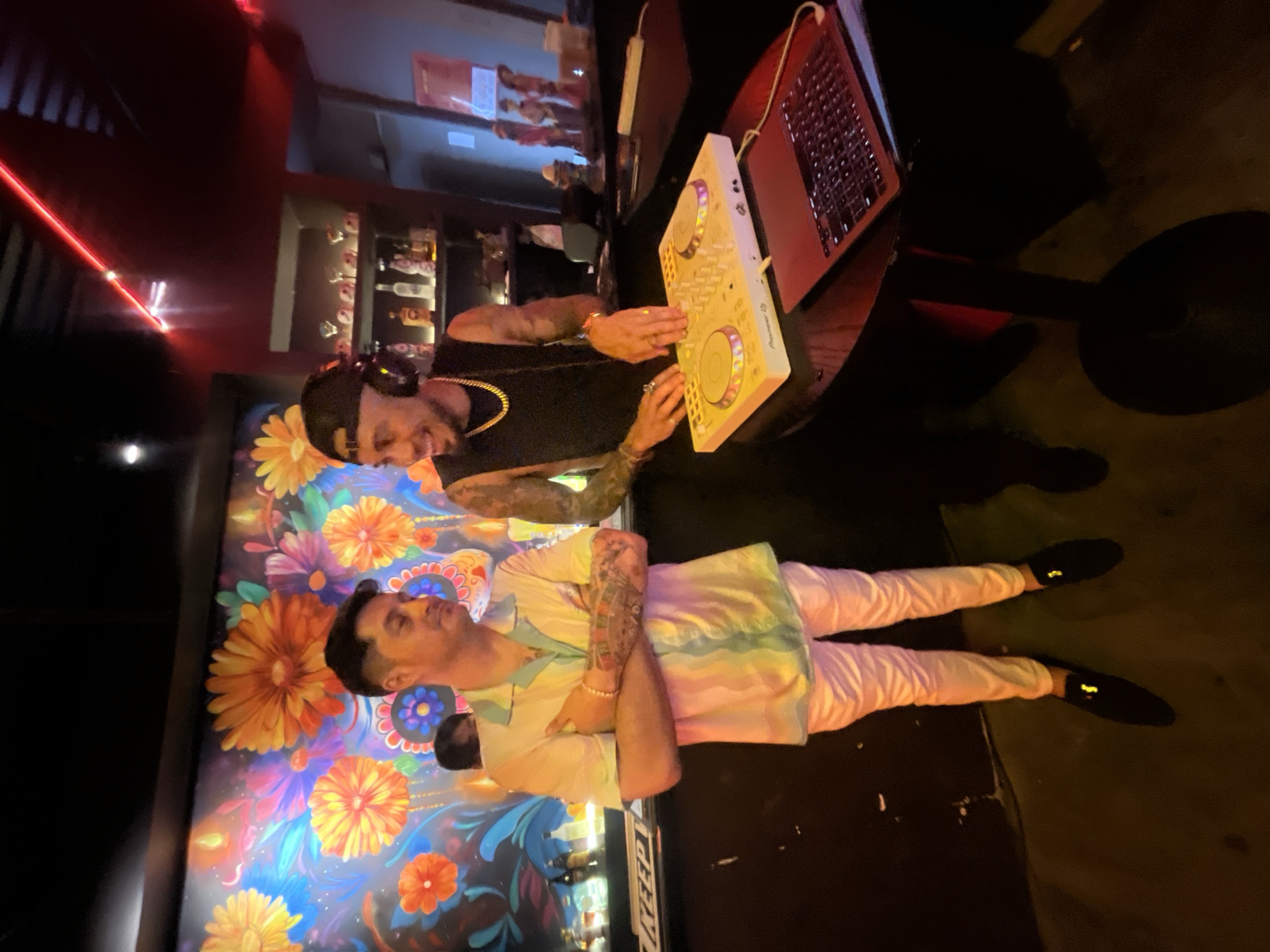 Two men are standing at a bar. The man on the left is wearing a light-colored shirt with pastel stripes and light pink pants. He has his arms crossed and is looking to his left. The man on the right is wearing a black tank top and a black baseball cap. He is wearing headphones and has his hands on a DJ controller. He is smiling and looking at the man on the left. There is a laptop on the table next to the DJ controller. In the background, there is a colorful mural of flowers. The setting appears to be a nightclub or bar. The image does not provide enough information to determine the specific location within the city of Đà Nẵng, Vietnam.