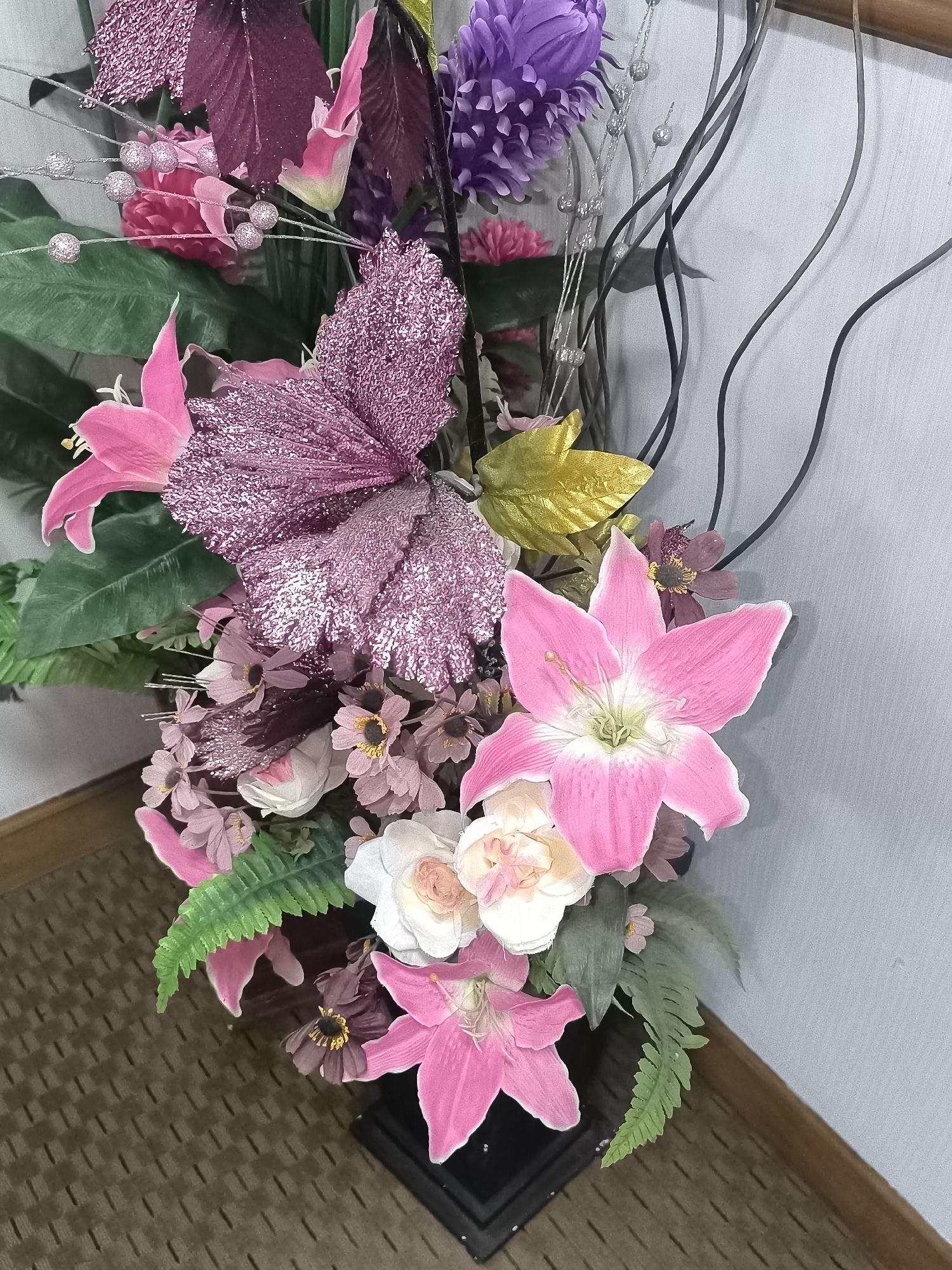 A tall, artificial floral arrangement with various pink, purple, and white flowers, accented with glittery pink butterflies and greenery, stands in a black vase. The arrangement is positioned in the corner of a room, with a wall visible behind it and a patterned carpet on the floor. The lighting is moderate, and the scene is static. There are no people present. The specific location in Kalabbirang, Indonesia is not discernible from the image.