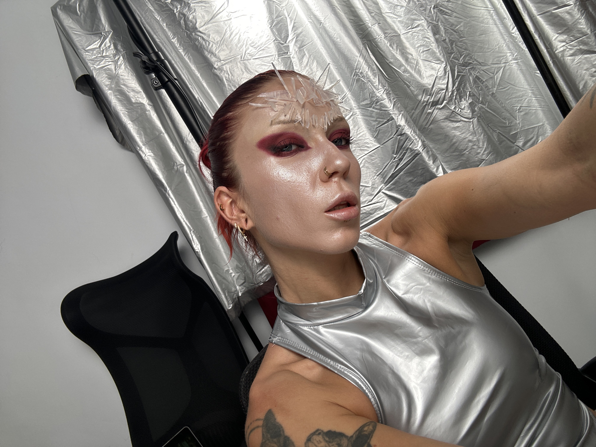 A person with red hair and bold makeup, adorned with a crystalline headpiece, is captured in a selfie. They are wearing a shimmering silver sleeveless top. The background consists of a crumpled silver fabric and a black object that appears to be a chair. The scene is likely indoors, with a plain white wall serving as a backdrop. The location in North Kuta, Indonesia, is not discernible from the visual information presented in the image.