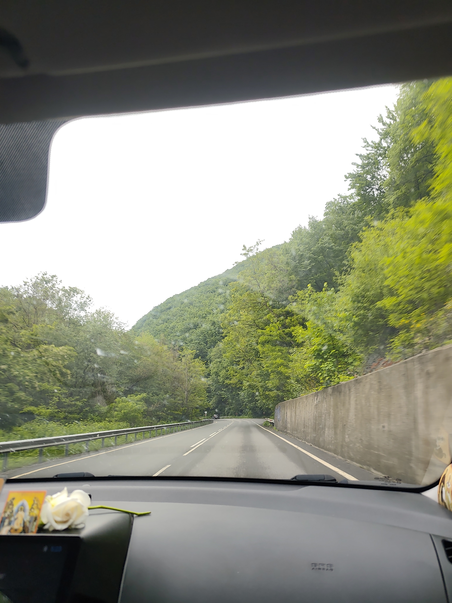 The image shows a view from inside a car driving on a paved road in a wooded area. The road is surrounded by trees and a concrete barrier. There is a small image and a white flower on the dashboard. The scene appears to be overcast. I'm unable to ascertain if this is the city of Dolno Osenovo, Bulgaria.