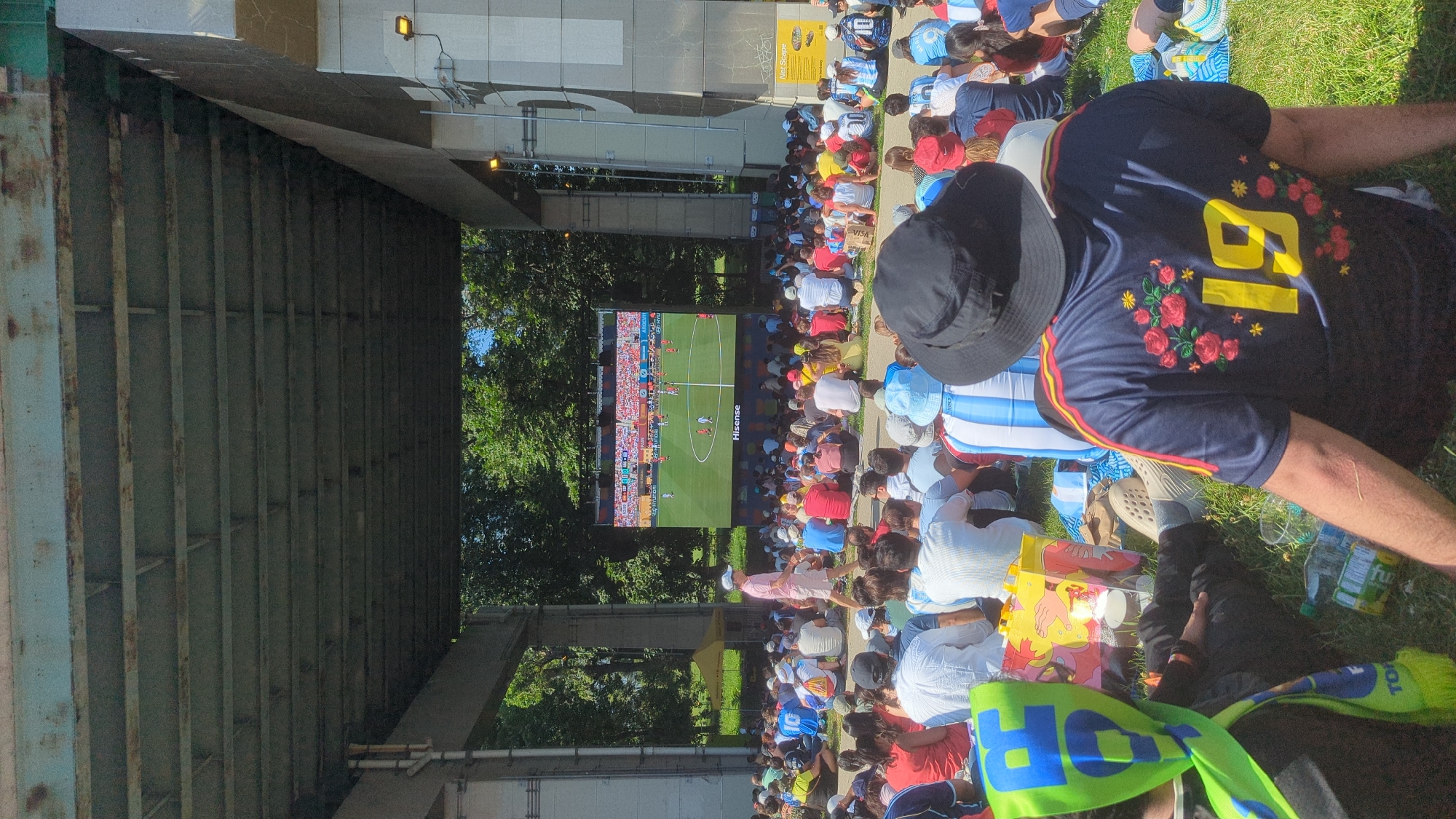 The image shows a large crowd of people sitting on grass, watching a soccer game on a big screen. The screen is placed under a bridge, and the people are spread out on the lawn, some sitting on blankets and others directly on the grass. Many people are wearing soccer jerseys, some with the colors of Argentina (blue and white stripes). In the foreground, a person in a black hat and a navy blue jersey with red roses and the number 19 is visible. The background shows trees and a yellow tent, suggesting an outdoor event. The event is likely taking place at Exhibition Place in Toronto, Canada, known for hosting large outdoor gatherings and watch parties for sporting events. The atmosphere appears relaxed and communal, with people of various ages enjoying the game together.