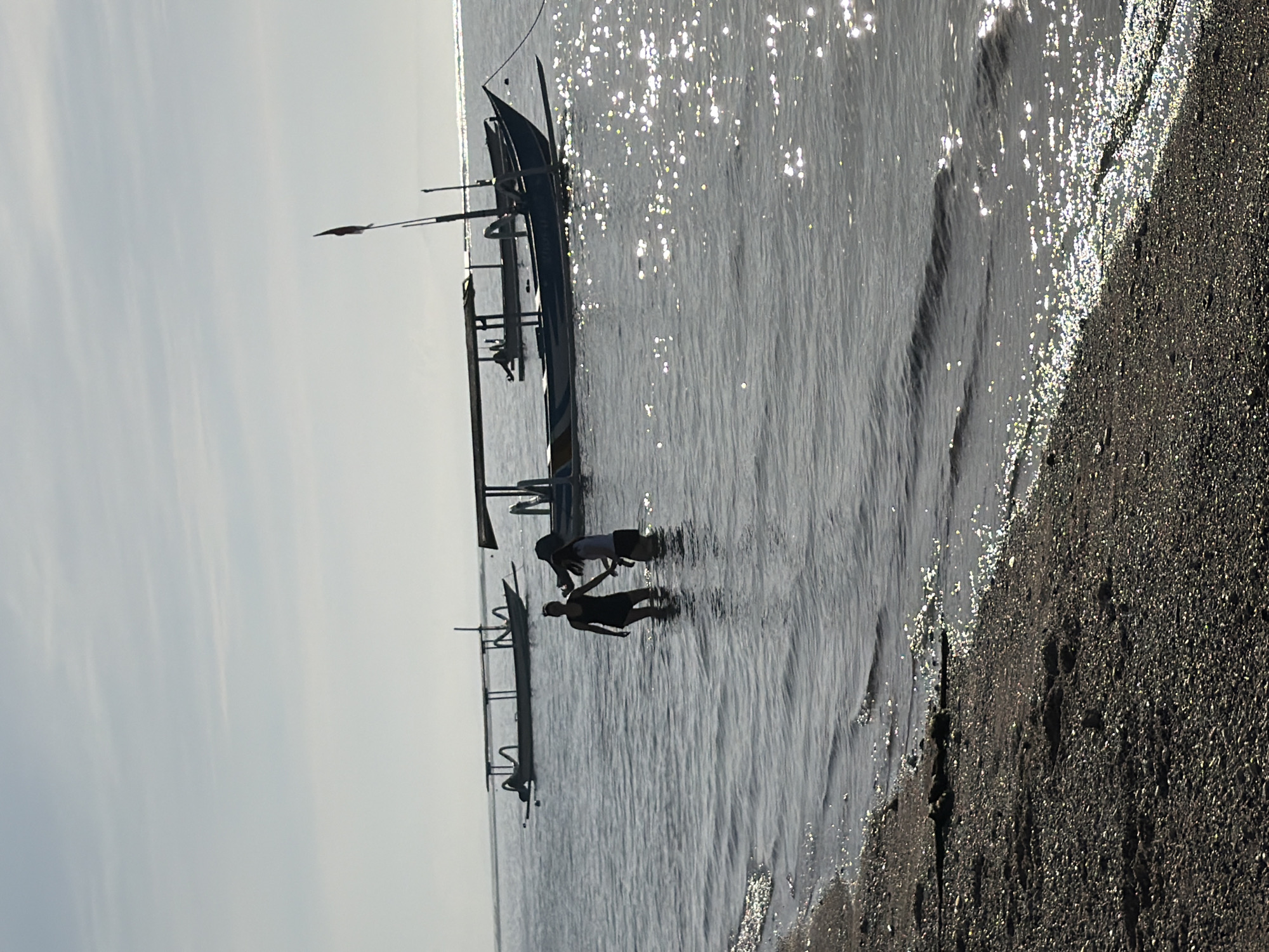 The image depicts a coastal scene in an unknown location in Indonesia, with the horizon line at the top. Dominating the scene is a row of traditional Indonesian outrigger boats, known as "jukung," anchored close to the shore. Two individuals are wading in the shallow water near one of the boats; their forms are silhouetted. The beach is composed of dark sand or pebbles. The water's surface reflects sunlight, creating shimmering patterns. The sky is overcast, indicating a hazy or cloudy day. The scene conveys a sense of daily life and maritime activity in a coastal community.