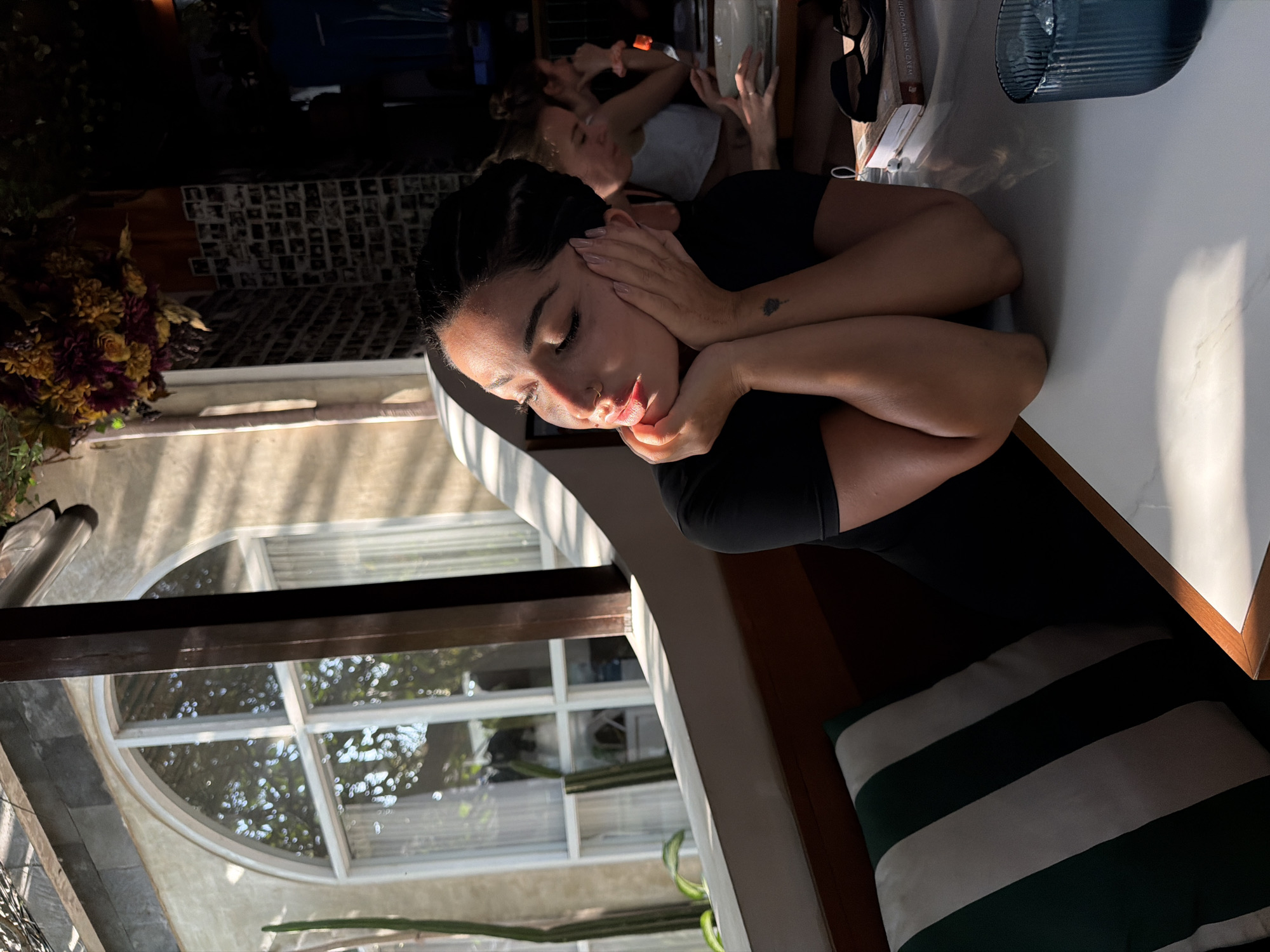 The image depicts a young woman with her hands on her cheeks, gazing to her left, illuminated by sunlight streaming through a window. She is seated at a table in a restaurant, evidenced by the presence of other patrons in the background, a vase of flowers, a book, sunglasses, and a glass of iced water. The restaurant appears to be in a tropical location, suggested by the lush greenery visible through the window and the architectural style of the building. The woman is wearing a black t-shirt and has her hair styled in braids. The scene captures a moment of quiet contemplation within a bustling urban environment.
