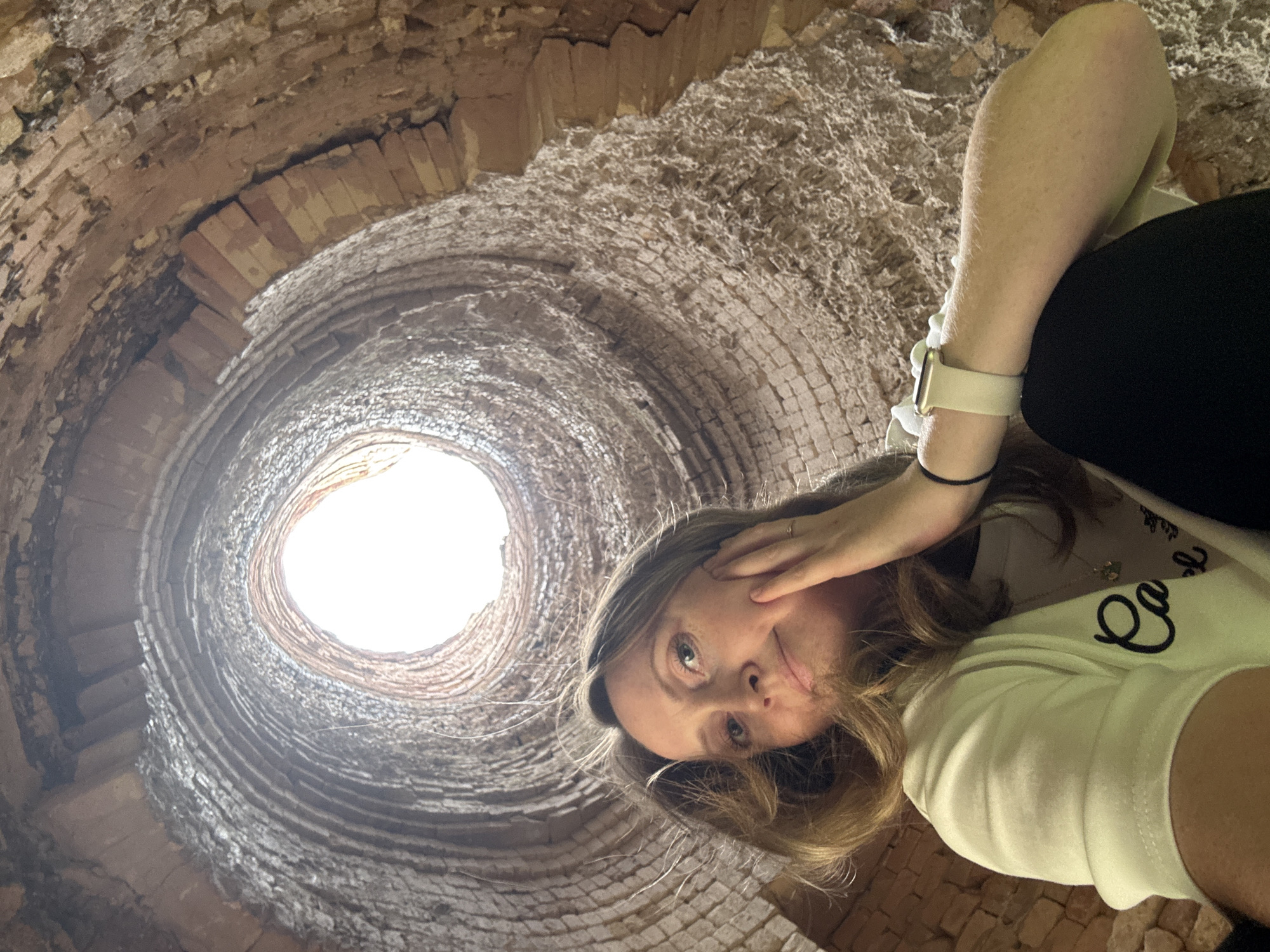 This is a low-angle, fisheye lens photograph of a young woman inside a circular, brick kiln or chimney structure. The subjects are the woman and the interior of the kiln. The woman is lying down, facing upwards towards the opening of the kiln, with her head tilted and resting on her hand, and her face is partially obscured by her hair. She is wearing a light-colored t-shirt with some text on it, and a white smartwatch. Her legs are visible in the foreground, and she is wearing dark pants.
The kiln is made of rough, weathered bricks, forming a cylindrical, tapering shaft. The bricks are of varying colors, from light brown to dark red, and some appear to be chipped or broken. The interior surface is textured and uneven. The opening at the top of the kiln is brightly lit by sunlight, suggesting it is daytime. There is no indication of the weather conditions.
The activity appears to be a selfie taken by the woman, posing within the unique architectural structure. The overall mood is casual and adventurous, with the woman seeming to explore or appreciate the unique environment. The location is identified as Mezhevoy, Russia, suggesting the kiln is a historical or industrial site in that area. Visible text on the woman's t-shirt is not legible.