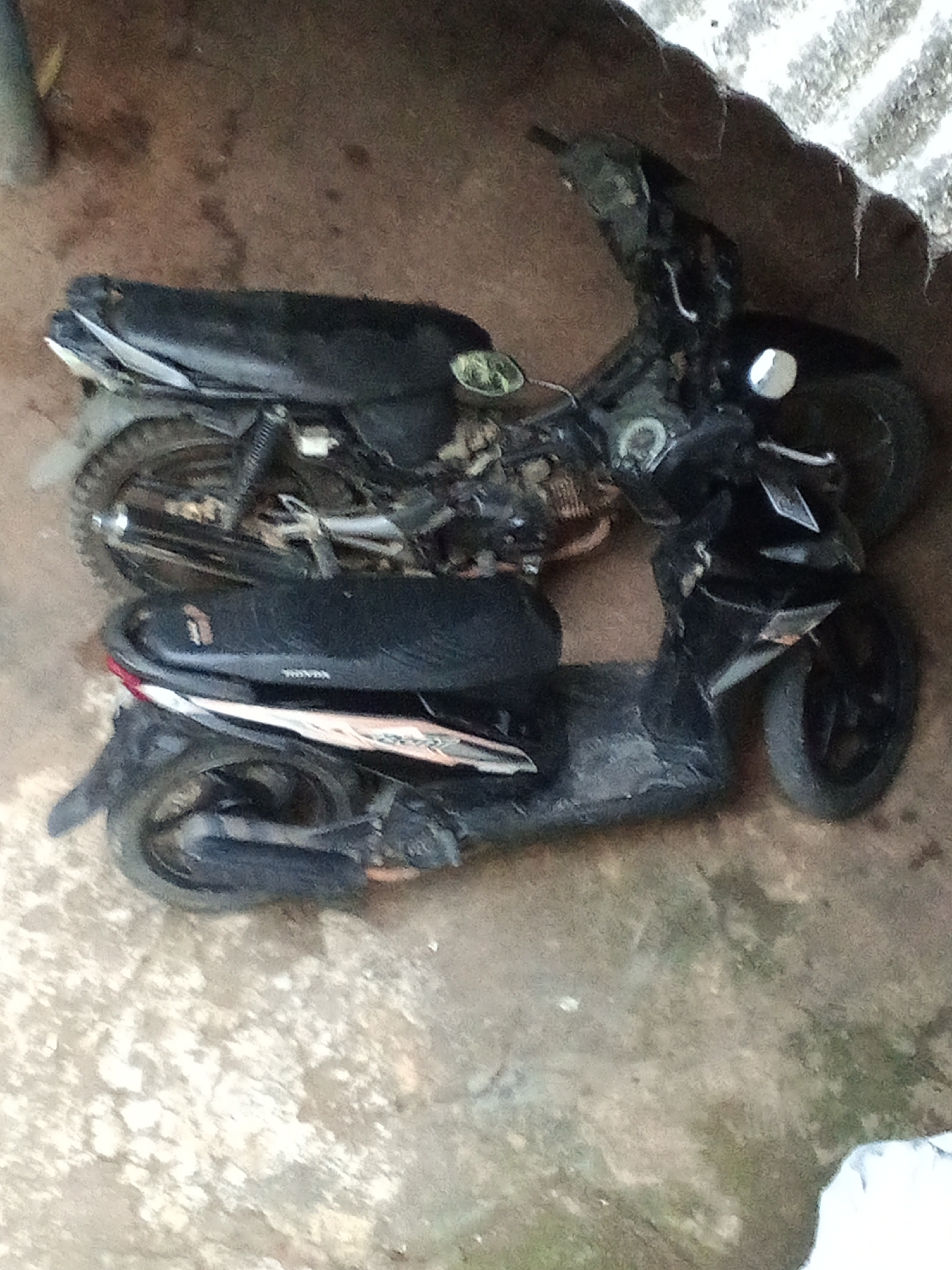 In Subang, Indonesia, two parked motorcycles are visible. The motorcycle in the foreground is a black scooter. The motorcycle in the background is a black motorcycle. They are parked on a dirt and concrete surface. The scene appears to be an outdoor area, possibly a yard or alleyway.