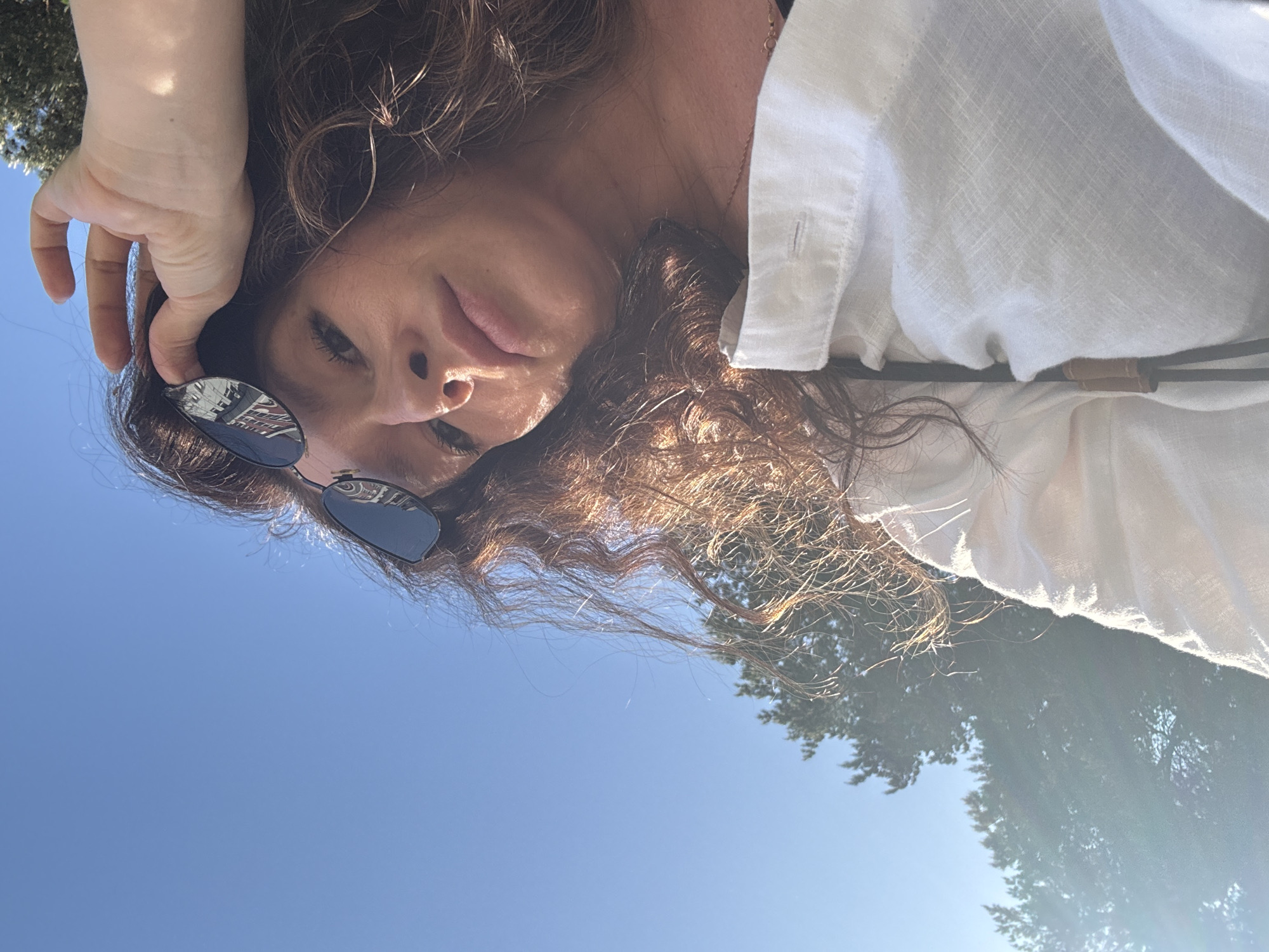 The image shows a woman wearing sunglasses and a white shirt lying on her back outdoors. The background consists of a clear blue sky and tree foliage, suggesting she is in a park or natural setting. Based on the location mentioned (Chaikovsky, Russia), the scene could be in a park or outdoor recreation area within that city. The woman's hand is near her head, possibly adjusting her sunglasses or hair.