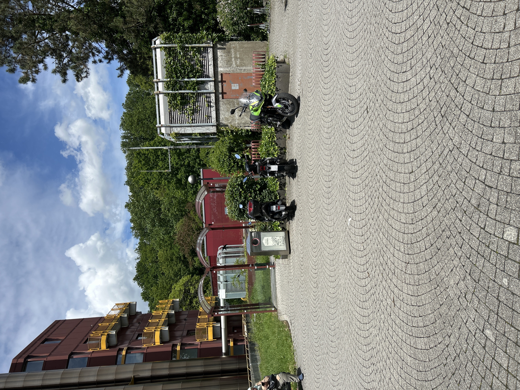 The image depicts an outdoor scene on a clear, sunny day in what appears to be a modern institutional or campus environment within Gummersbach, Germany.
In the foreground, a large paved area made of light-grey cobblestones is visible, laid in a pattern of concentric arcs. Three motorcycles are parked on this paved surface. On the far left, a dark-colored scooter with a German license plate "087 HSK" is parked, facing away from the viewer. To its right, another dark-colored motorcycle is parked facing away. On the right, a prominent green and silver sport-touring motorcycle is parked, facing the viewer.
To the left of the paved area, a grassy strip borders a silver bench. A single individual is seated on this bench, looking down at a mobile device held in their hands. They are wearing light-colored clothing and blue footwear, with a bag next to them.
In the mid-ground, a multi-story building with reddish-brown cladding and numerous rectangular windows is situated on the left. It features distinctive bright yellow cantilevered balconies with railings. A large, cylindrical, metallic pipe runs vertically along the building's left side. Adjacent to the grassy area, a dark grey trash receptacle with "REST MÜLL" (general waste) indicated on one compartment stands near two arched, red-framed shelters with transparent roofs.
Further right, a series of industrial-style concrete structures are present. The most prominent is a concrete building partially covered in green ivy, featuring louvered vents and a rusty-red door. Metal framework extends above this structure. In front of it, red metal railings are installed. To the left of this building, a tall metal pole with horizontal cross-elements, possibly an antenna array or weather instrumentation, stands against a backdrop of dense green trees. Another lamppost with a spherical light fixture is also visible. The background is dominated by a lush, dense forest under a blue sky with scattered white cumulus clouds.