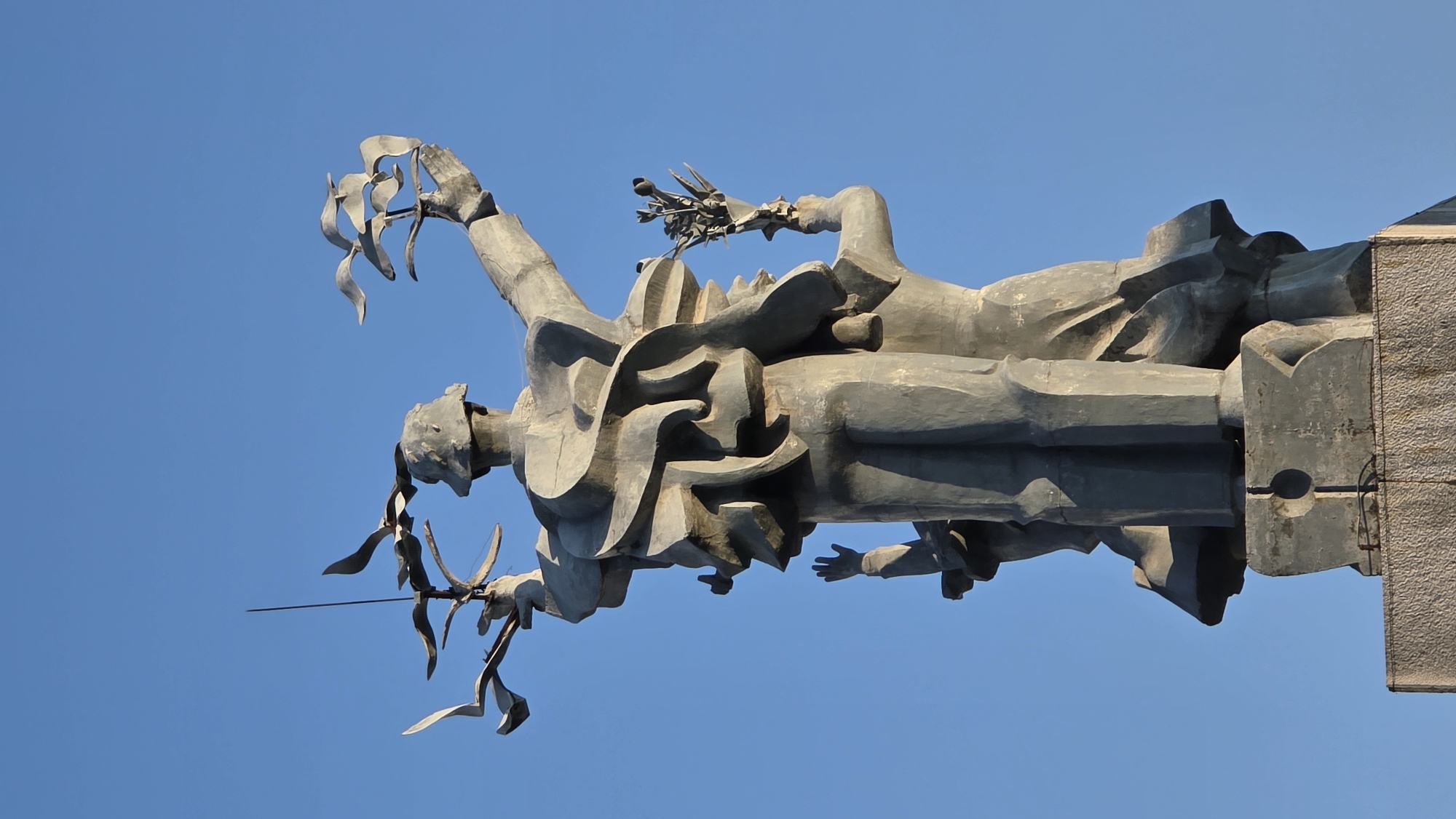 The image shows a close-up, low-angle view of a monument against a clear blue sky. The monument appears to depict two figures, possibly a male and female, both rendered in a stylized, angular, gray concrete or metal material. The male figure on the left holds a sword or similar weapon adorned with abstract metal appendages resembling flames or wings. The female figure on the right carries what appears to be a sheaf of wheat or olive branches. The figures stand attached to a concrete plinth or base on the right. This statue is the Monument to the soldiers who died for the country in the city of Nha Trang, Vietnam.