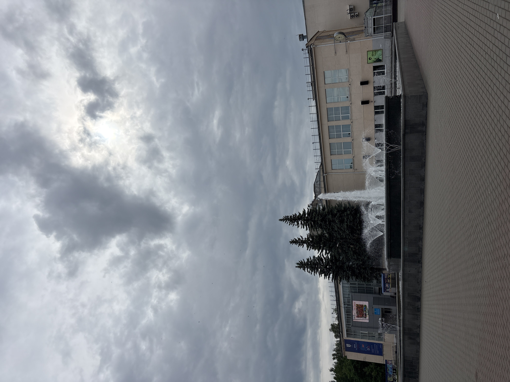 A medium shot depicts a cityscape, likely in the city of Chaikovsky, Russia, featuring a gray sky filled with clouds, partly obscuring the sun. Below, there are buildings, three fir trees, and a multi-tiered fountain with water spouting. The building nearest to the fountain has multiple windows and an exterior clock. The fountain itself has multiple levels of water flowing, suggesting an architectural design. A section of cobblestone pavement is on the right of the frame. No people are visible.